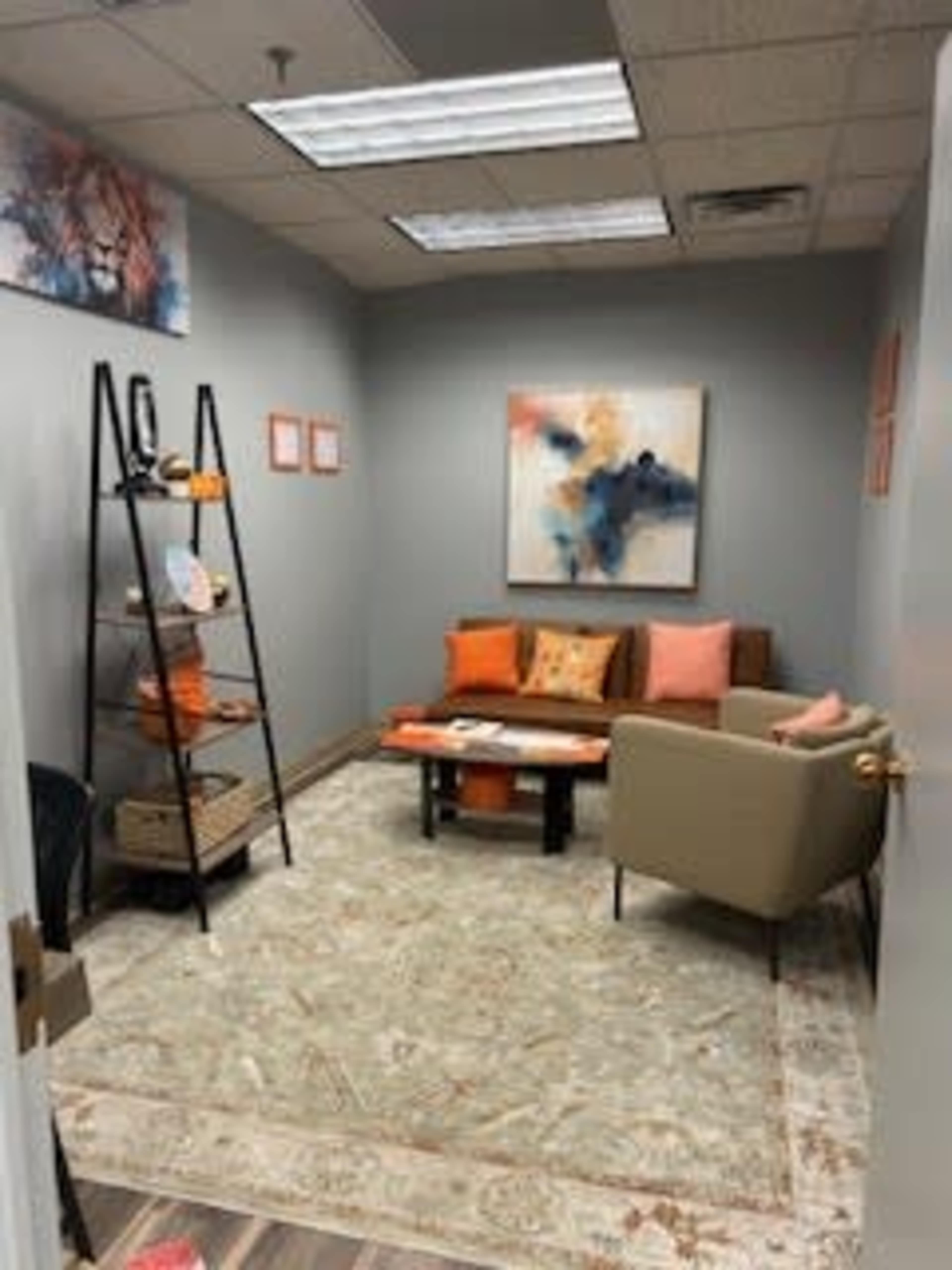 The image shows a small office space featuring a seating area with a couch, an armchair, and a coffee table, along with a decorative rug and shelves displaying colorful items on the walls.