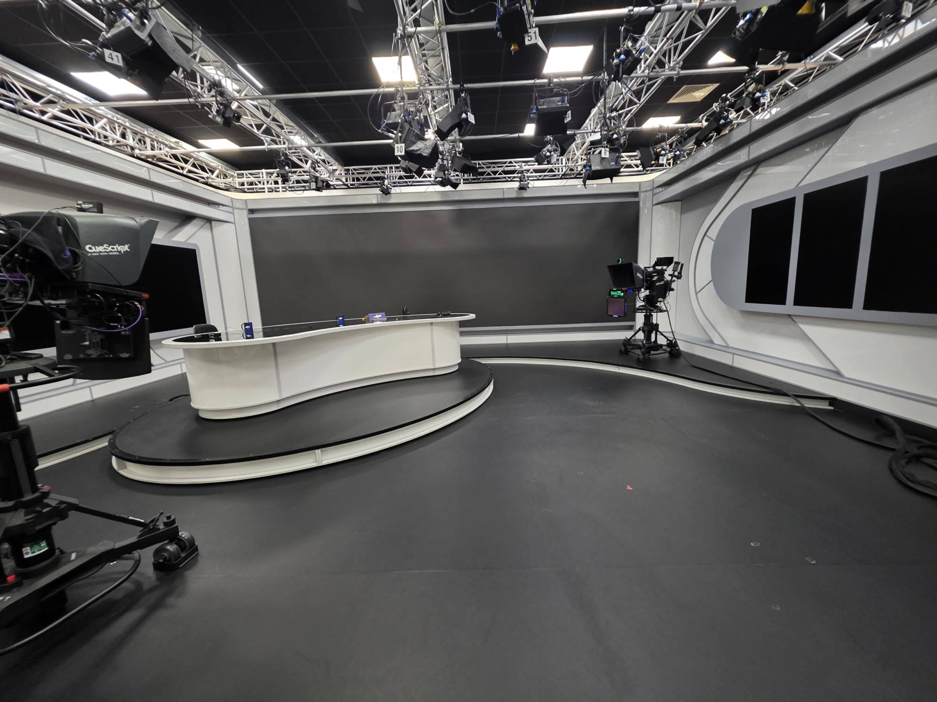 The image shows a television studio with a modern design, featuring a curved white news desk, multiple cameras, and a dark background.