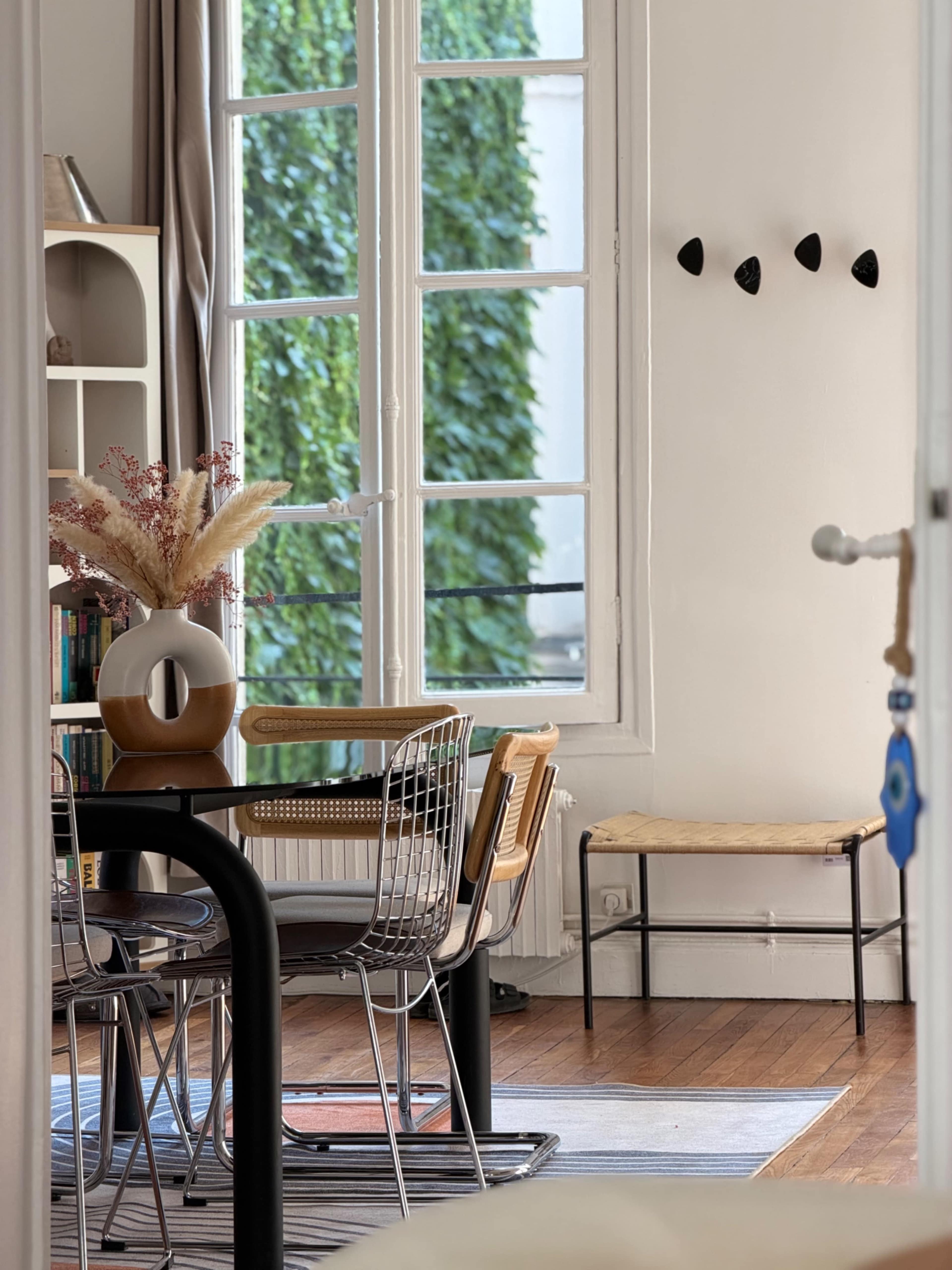 A modern dining area features a round black table with metal chairs, a decorative vase, and large windows allowing natural light to illuminate the space.