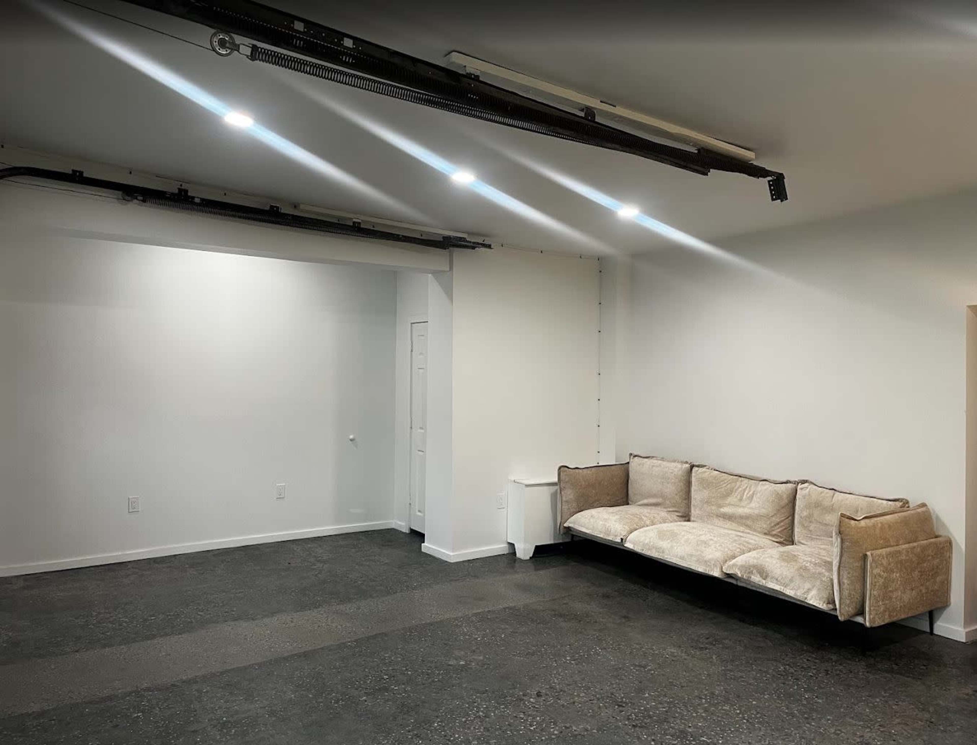 The image shows a spacious, minimalist room with concrete flooring, a white wall, and a light-colored couch placed against one side.