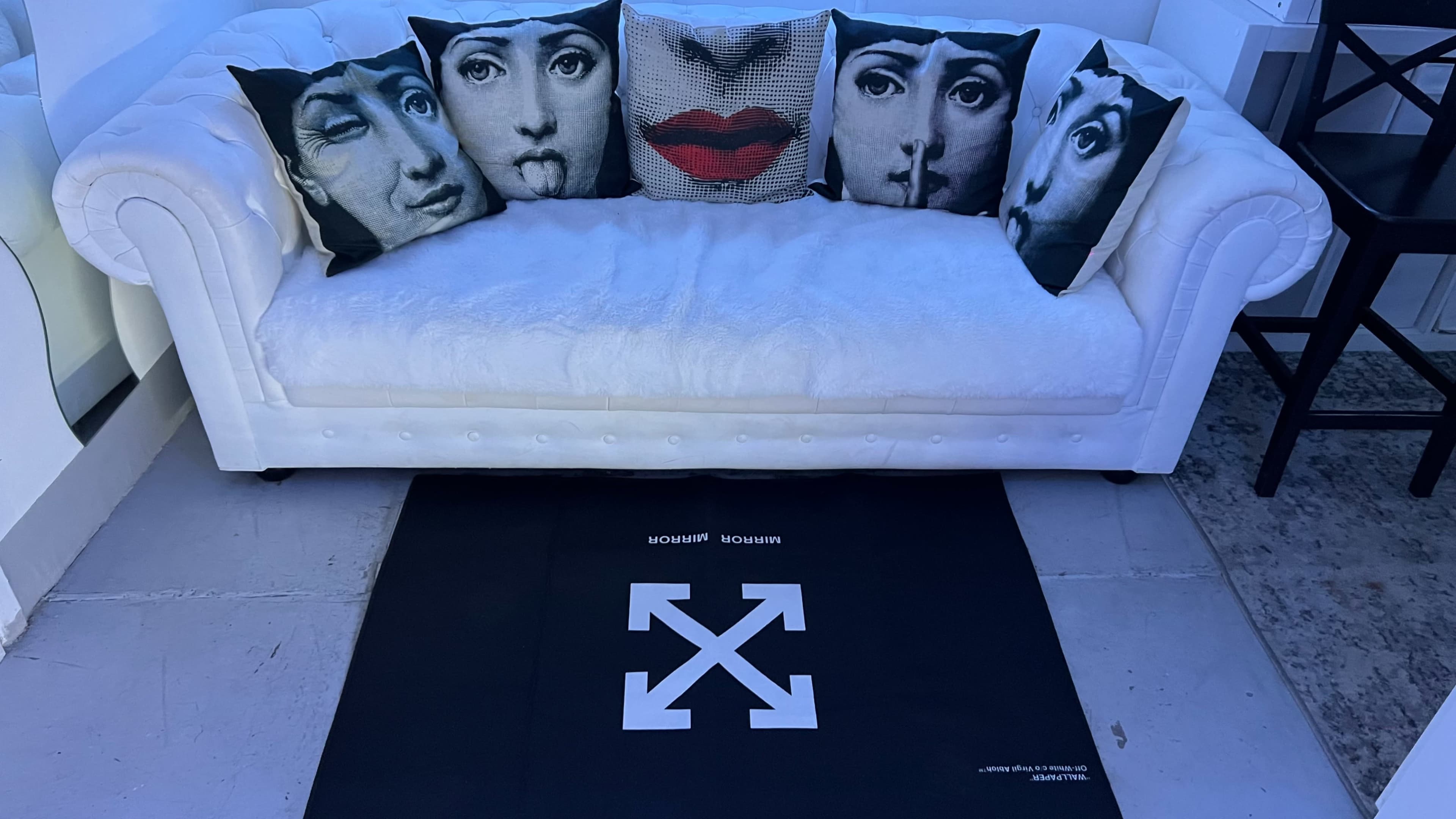 A white couch with decorative pillows featuring various facial expressions is placed on a black mat with a distinctive logo.
