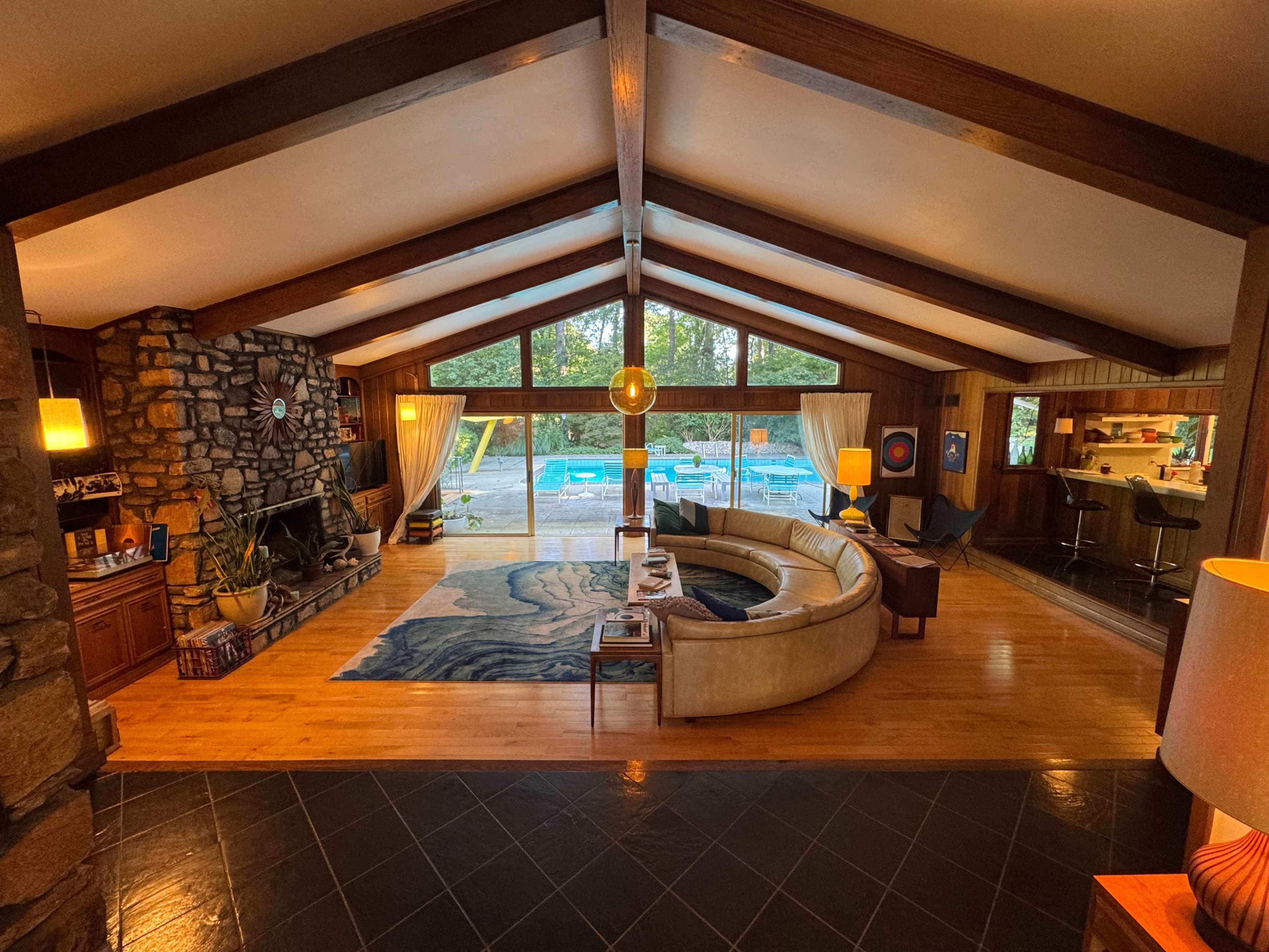 The image shows a spacious living room with a curved sofa, a stone fireplace, large windows overlooking a pool area, and wooden beams on the ceiling.