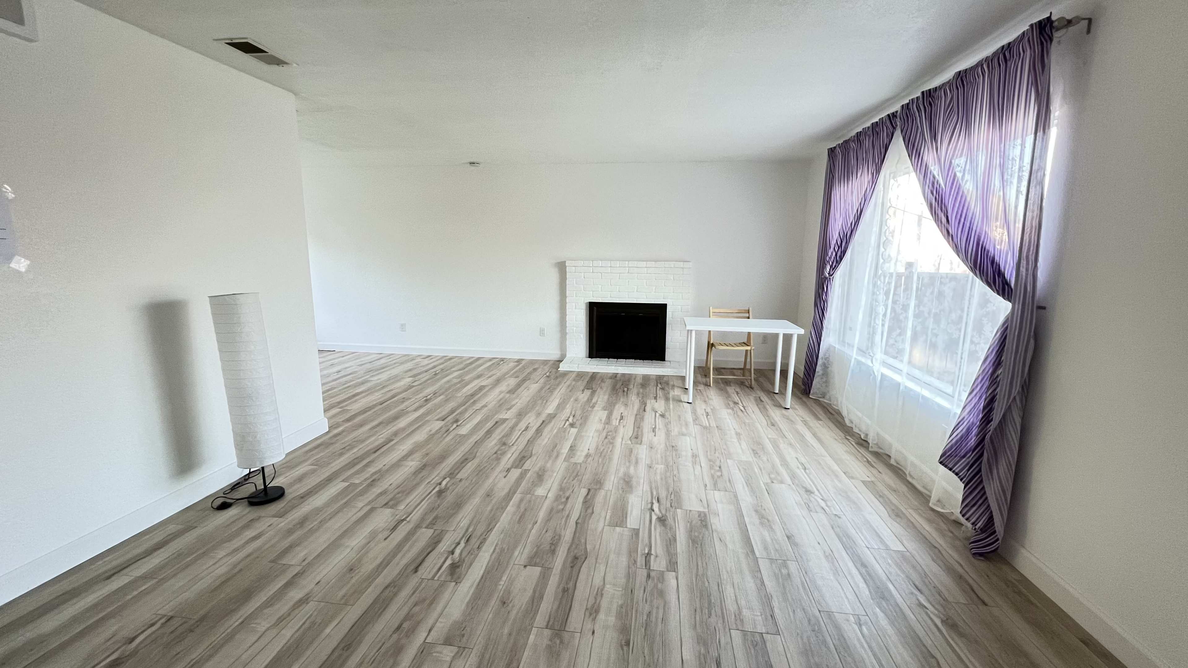 A spacious, unfurnished living room features a white fireplace, a small table with a chair, large windows with sheer curtains, and light-colored flooring.
