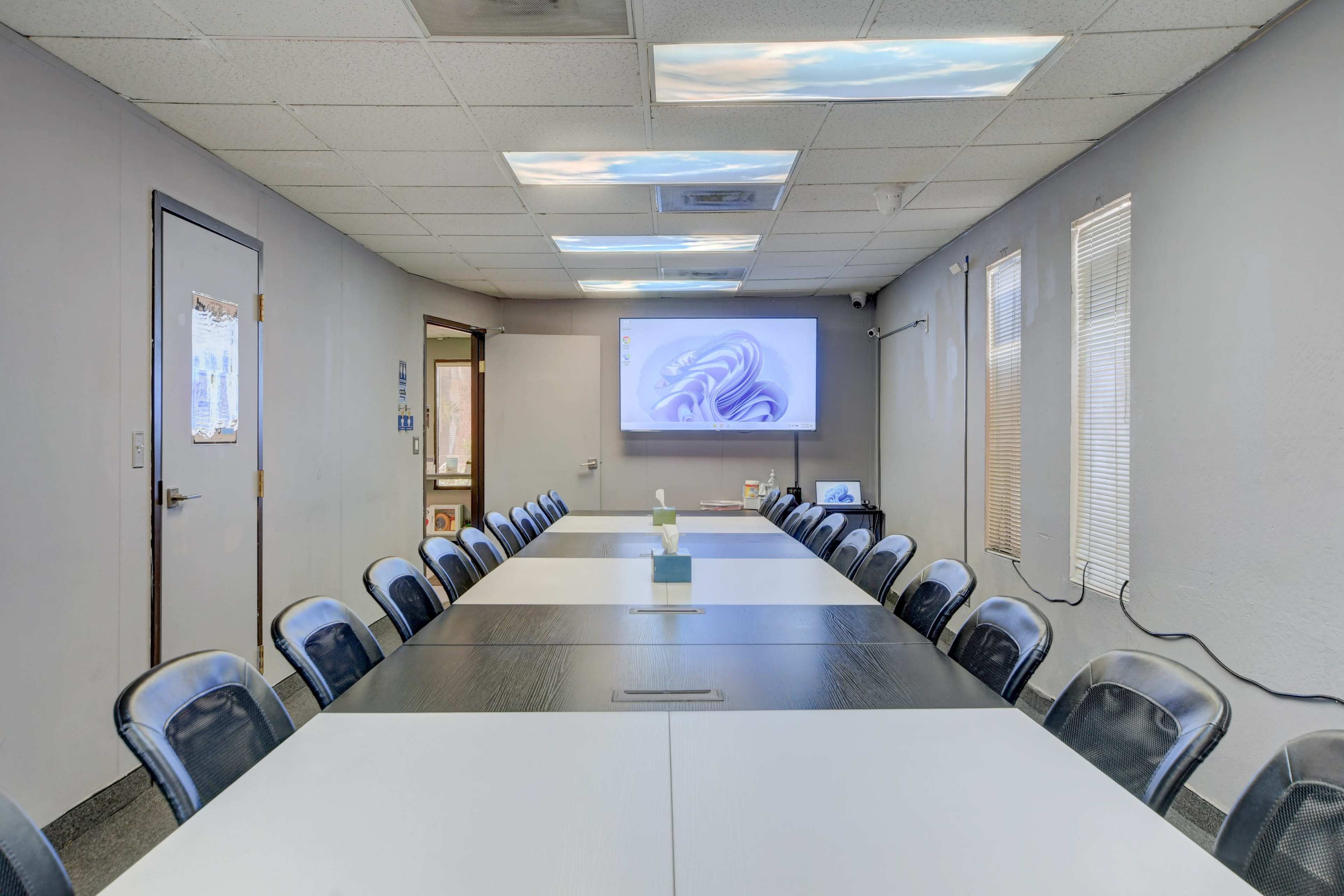 The image shows a conference room with a long table, black chairs, and a large screen displaying a presentation on the wall.
