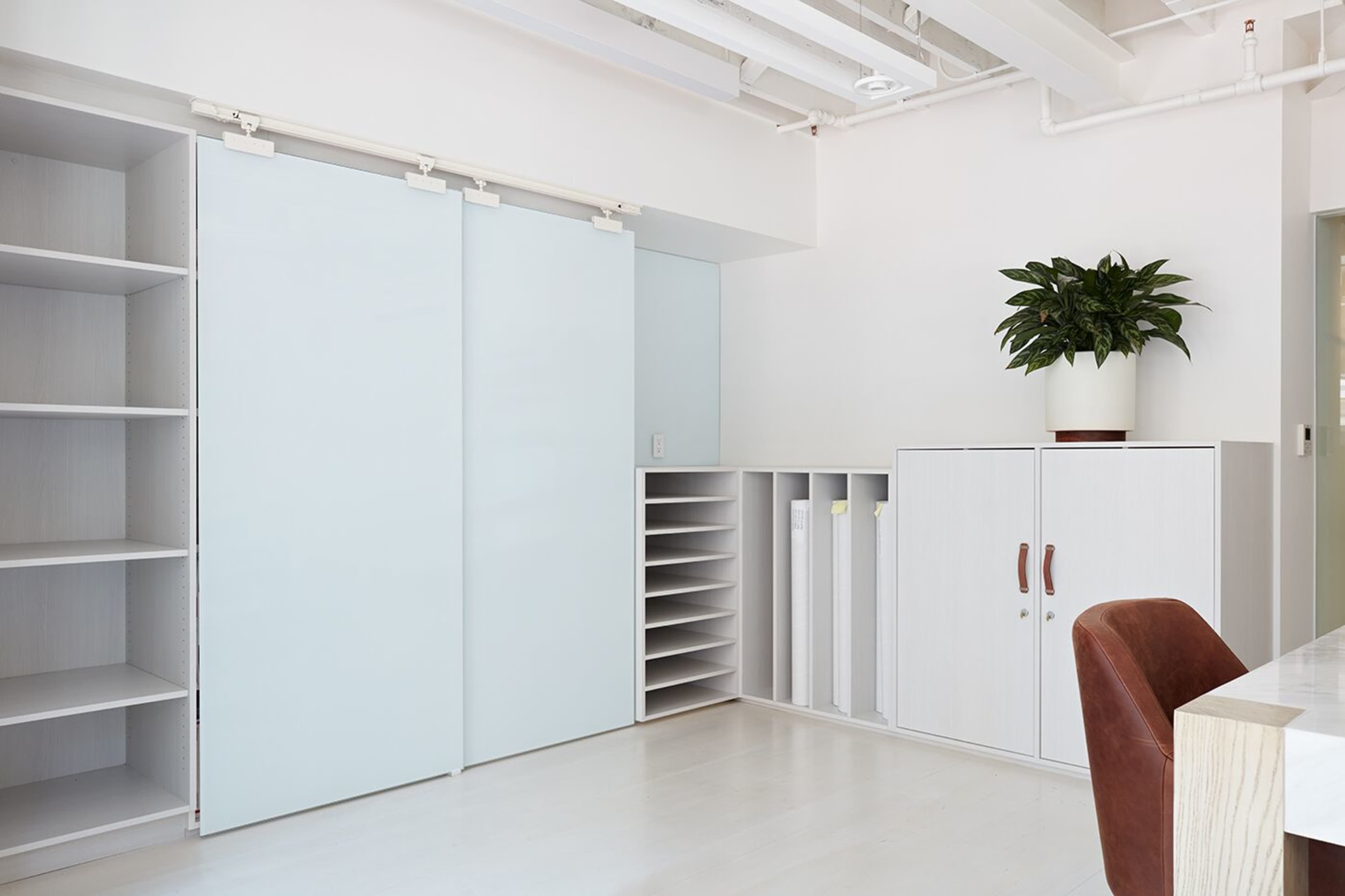 A modern, minimalist office space with sliding doors, shelving units, and a potted plant.