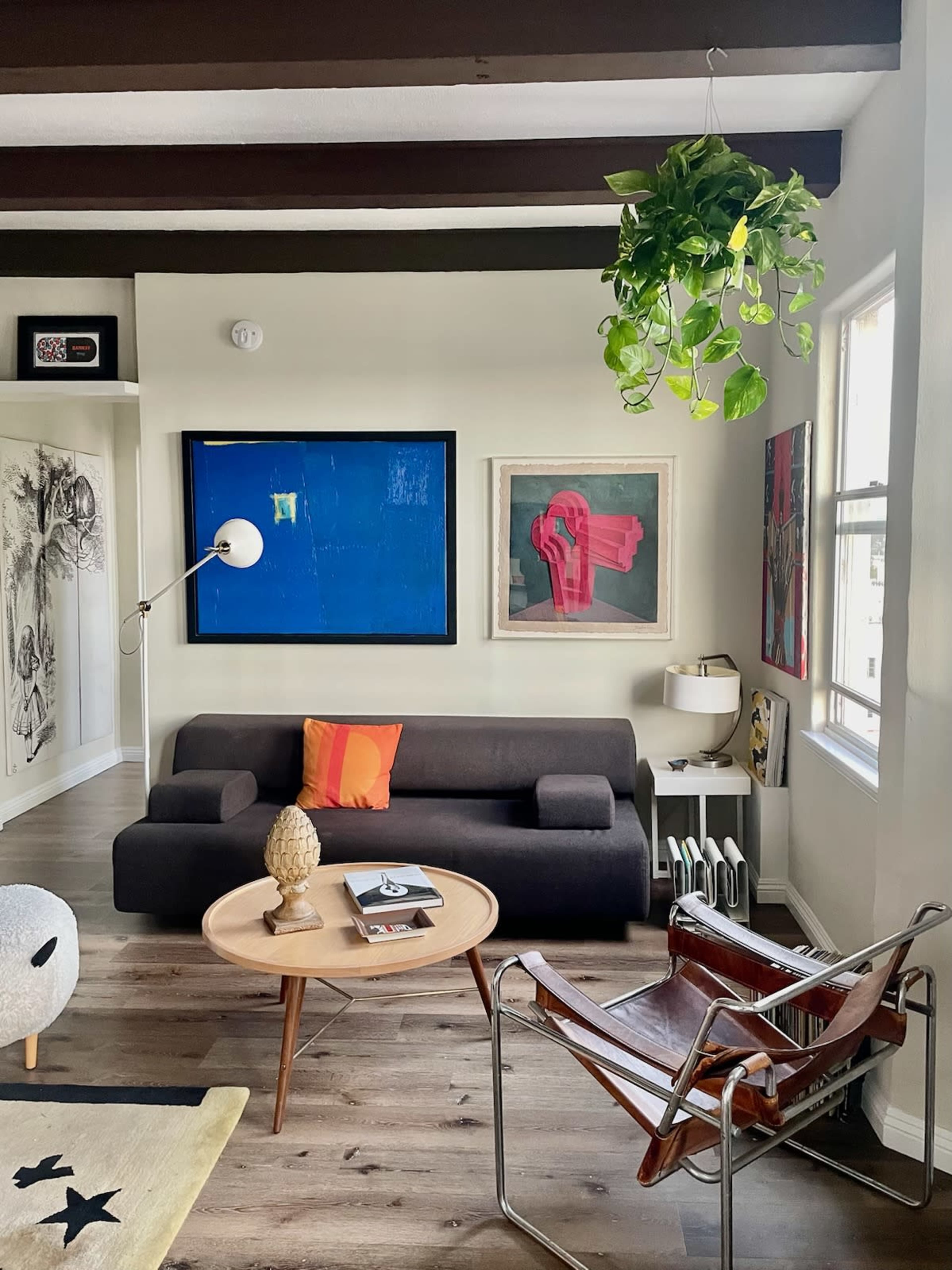 A modern living room featuring a dark sofa, a round coffee table with books, and art pieces on the walls, including a blue painting and a plant hanging from the ceiling.
