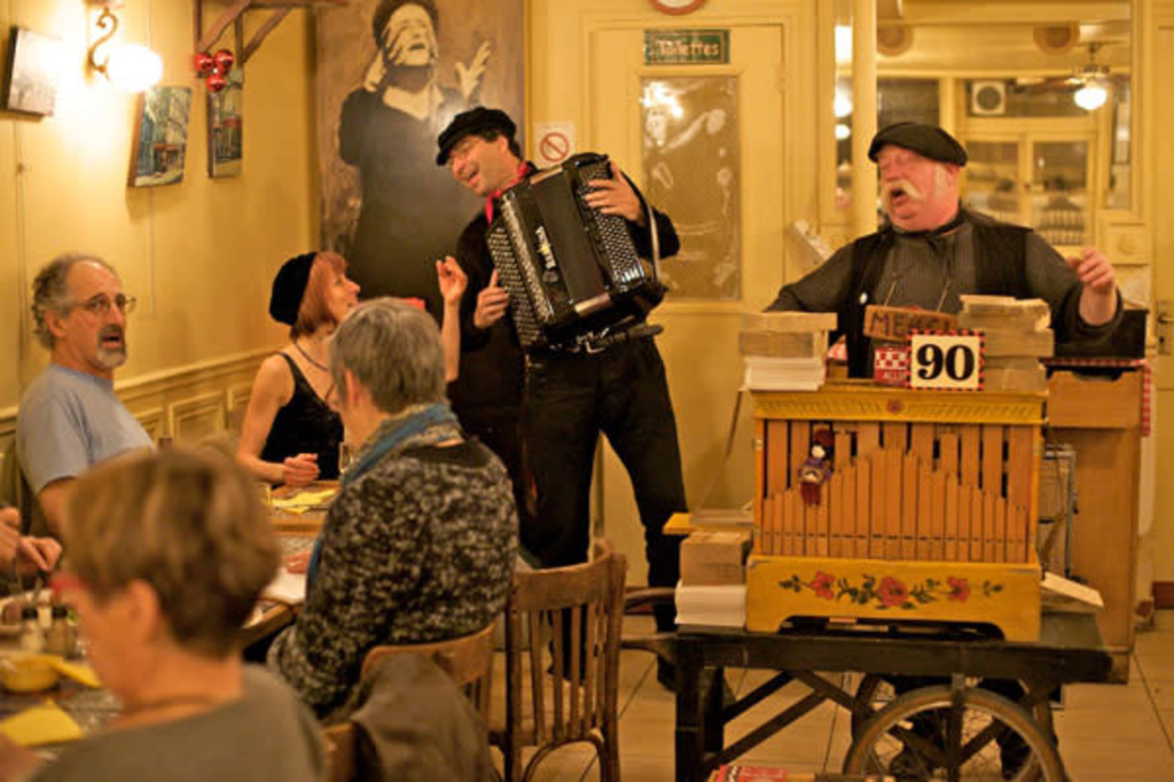 Musicians perform with an accordion and a street organ while diners enjoy their meals in a café setting.