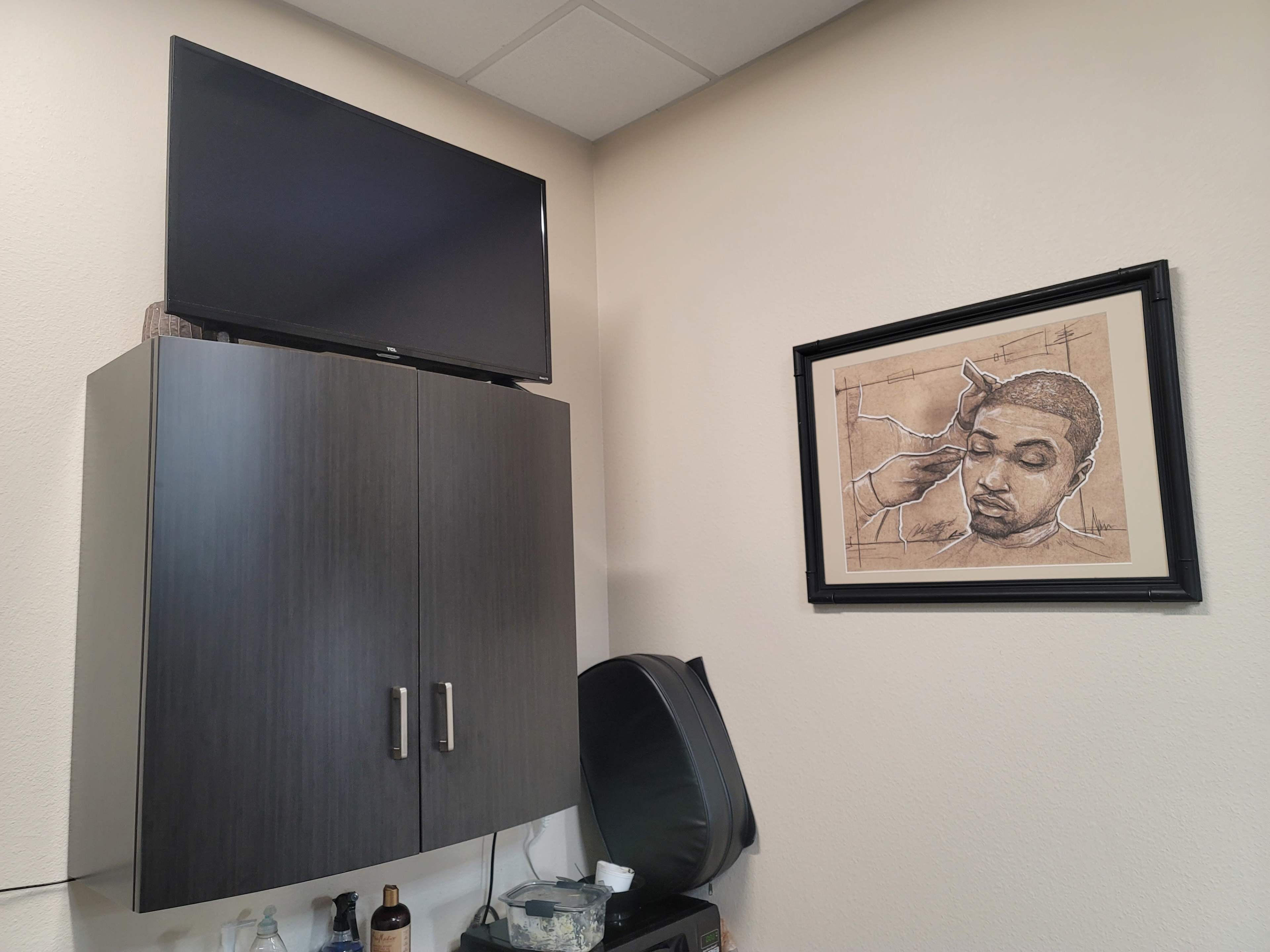 A wall with a mounted television, a framed artwork of a person getting a haircut, and a dark cabinet beneath them.
