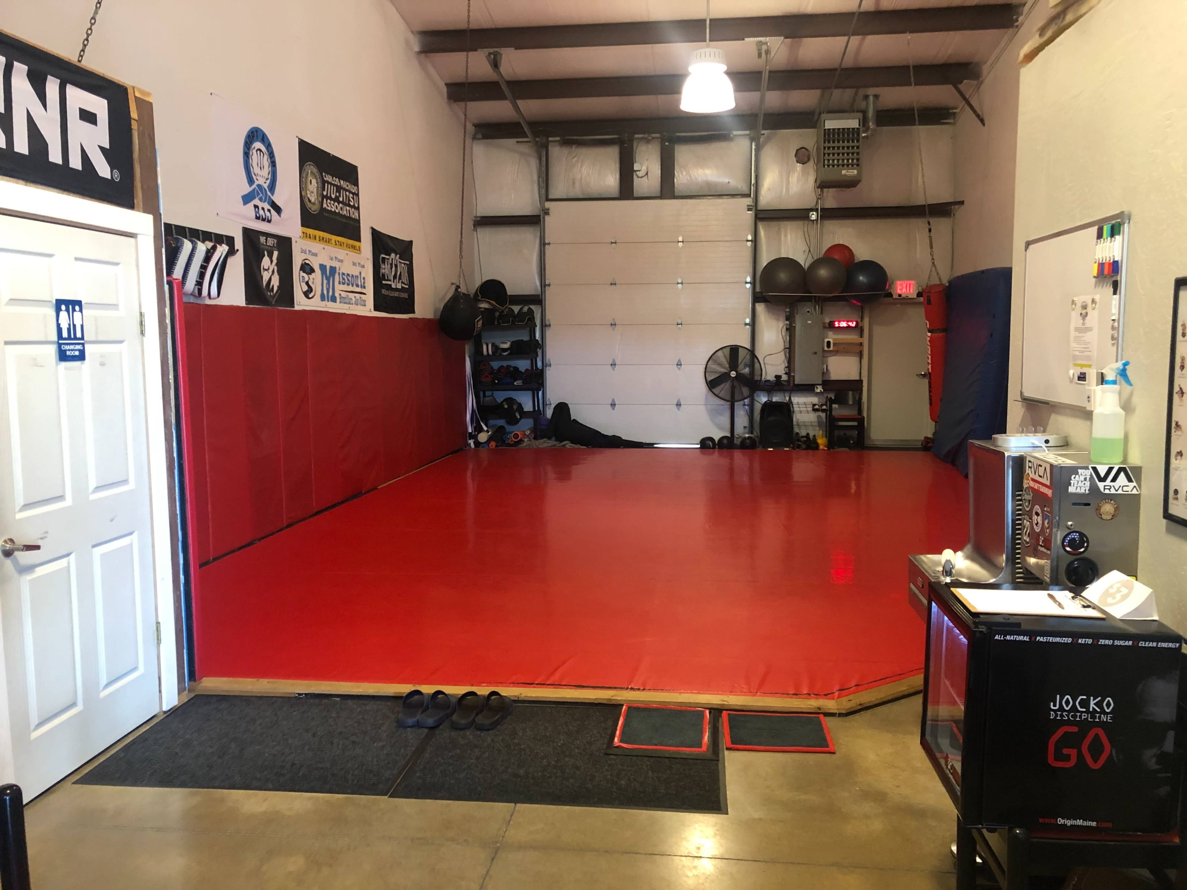 A martial arts training facility with a red mat, gym equipment along the walls, and a door leading to a restroom.