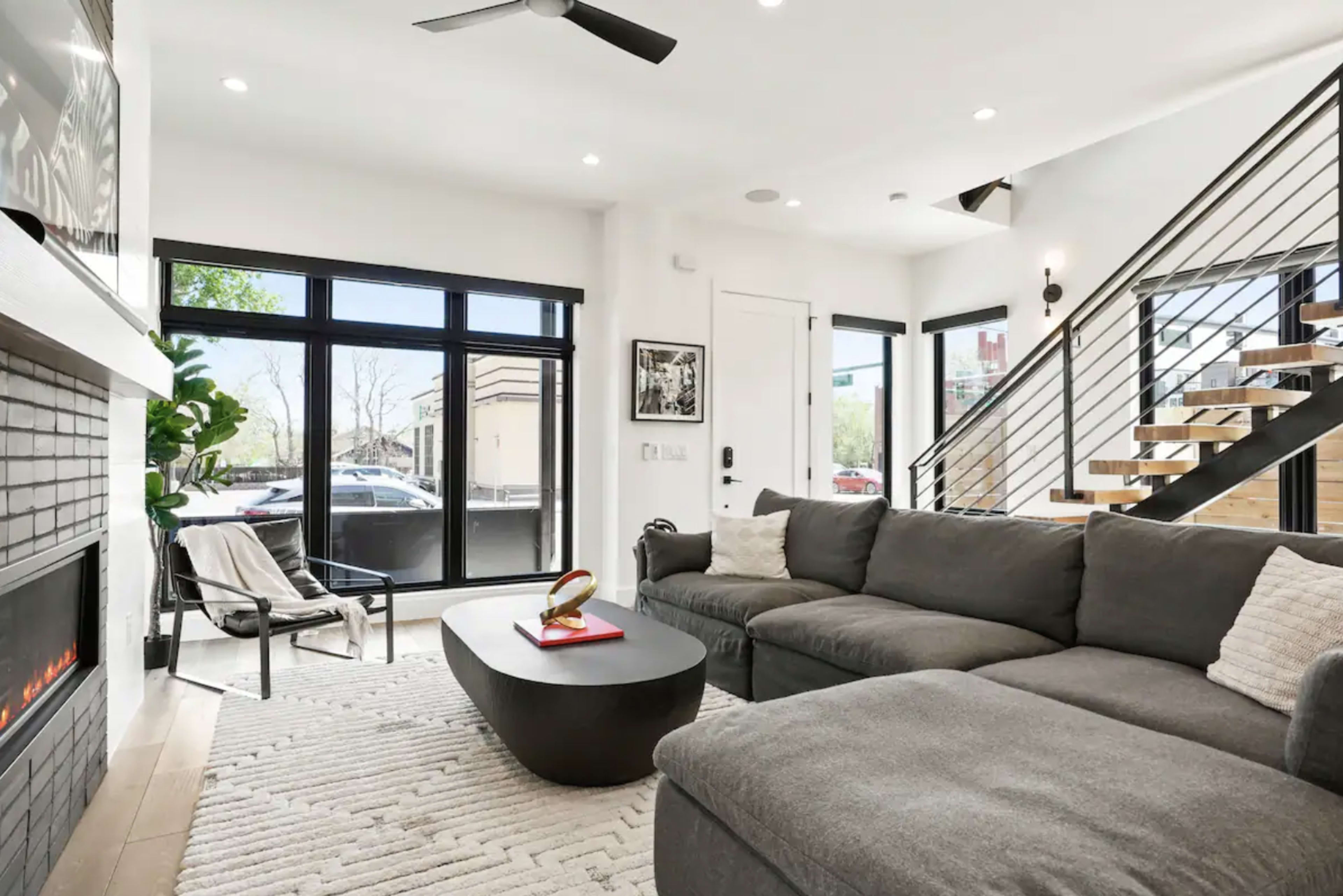 A modern living room features a gray sectional sofa, a black coffee table, and large windows with natural light, alongside a staircase leading to the upper floor.