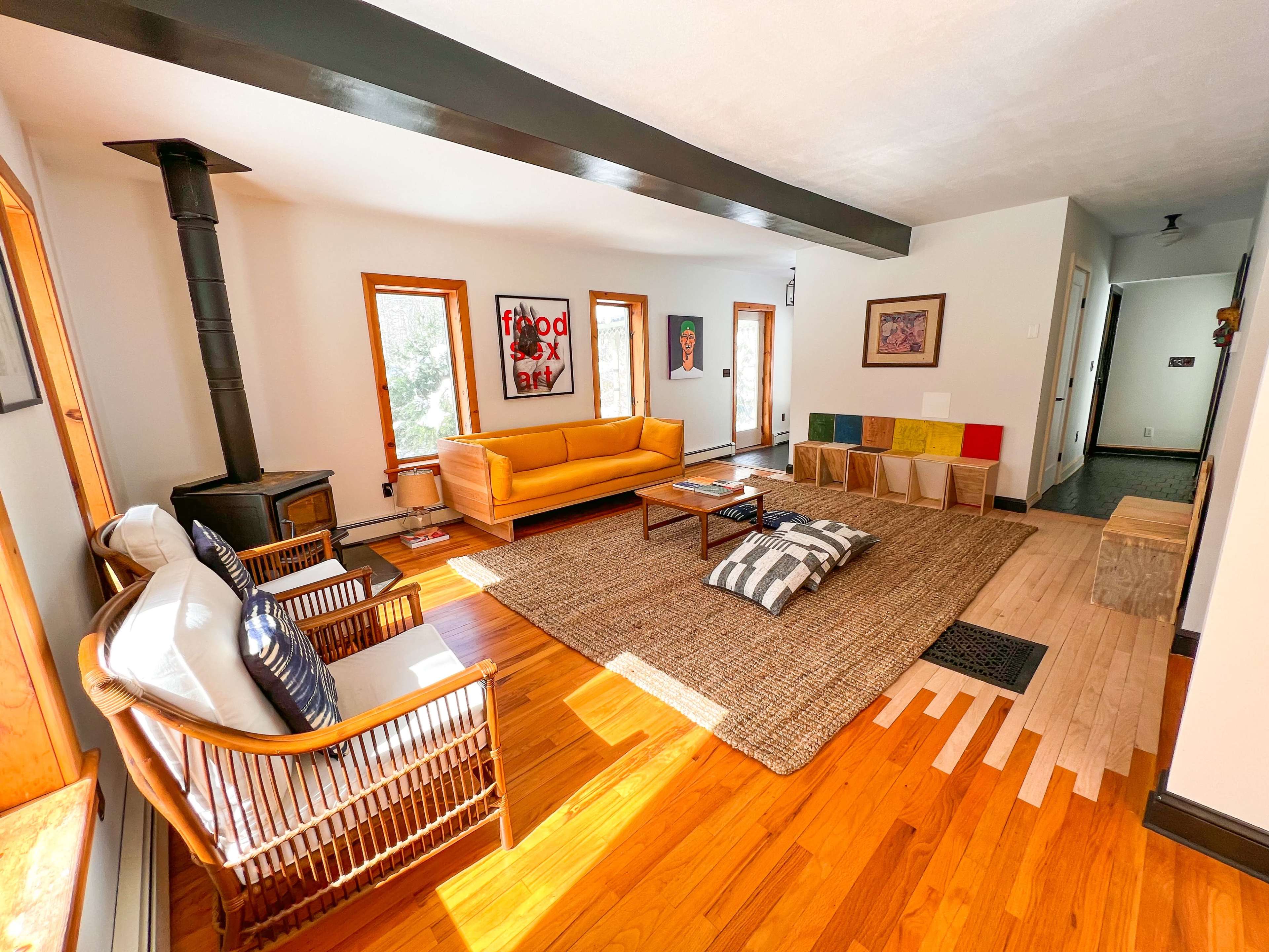 A spacious living room with a yellow sofa, a wood-burning stove, and a rug surrounded by several cushions and wooden decor.