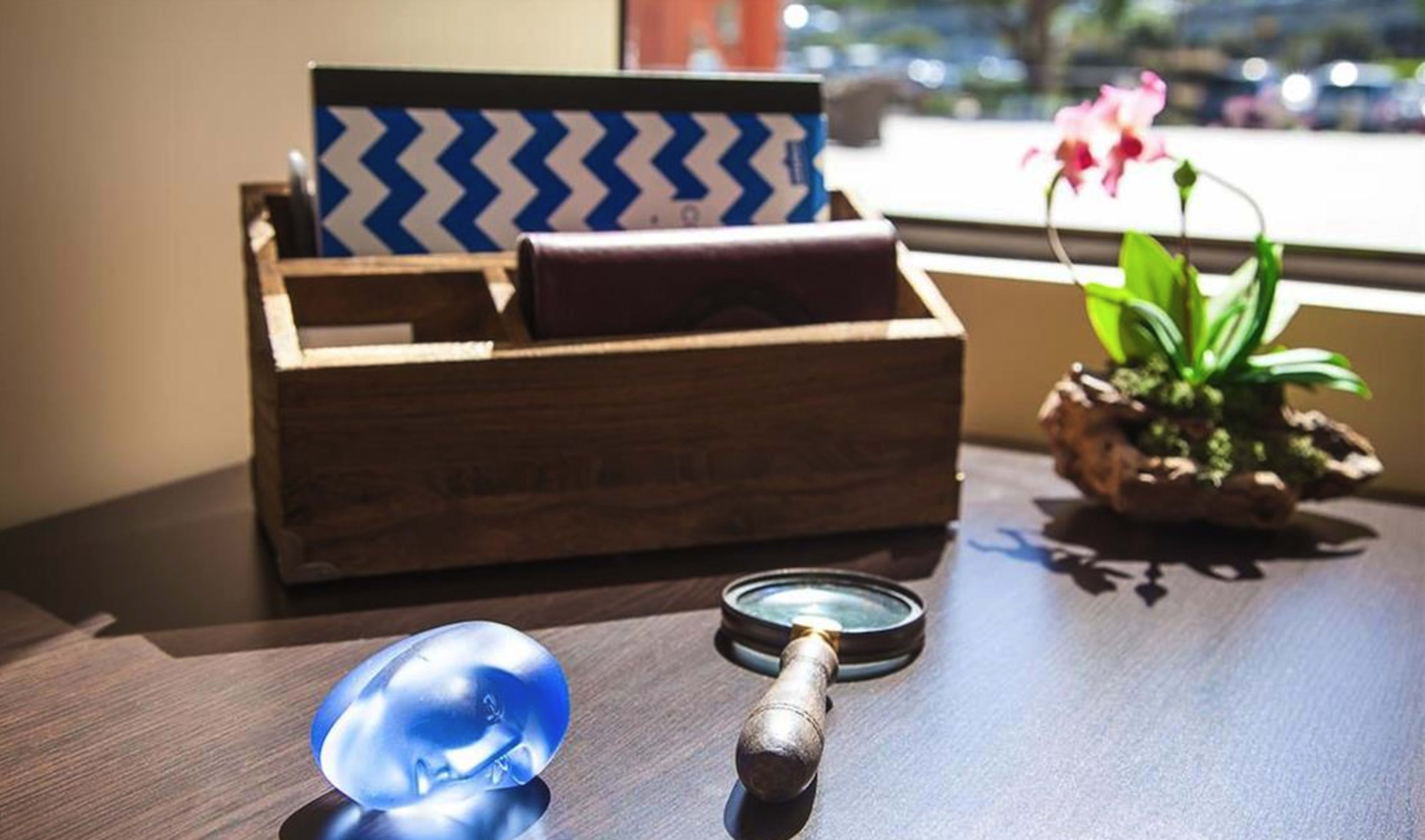 A wooden organizer holds files and a wallet on a table, alongside a blue glass object and a magnifying glass near a small potted orchid.