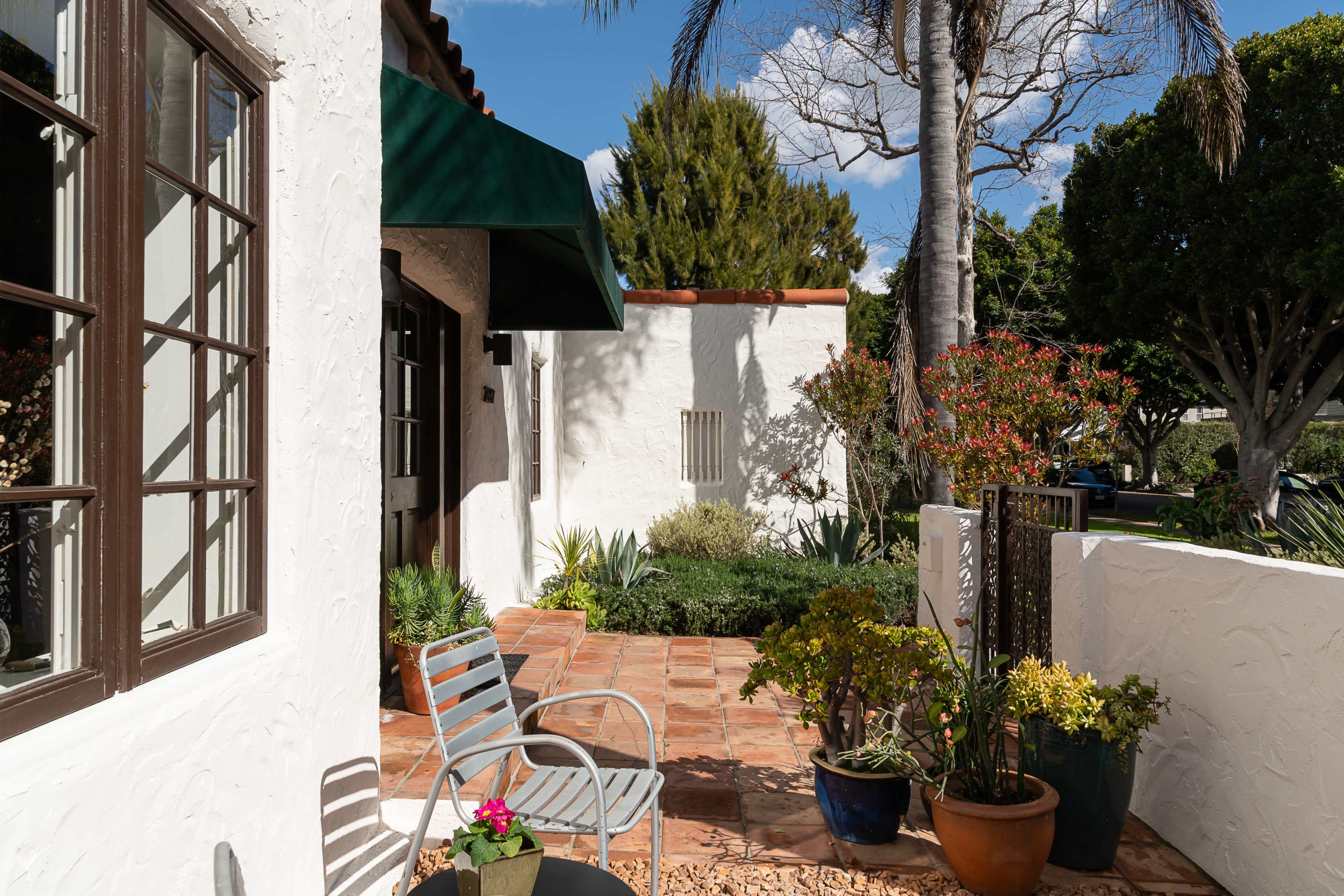 A sunlit patio with potted plants, a stone path, and a chair beside a house featuring textured white walls and green awnings.
