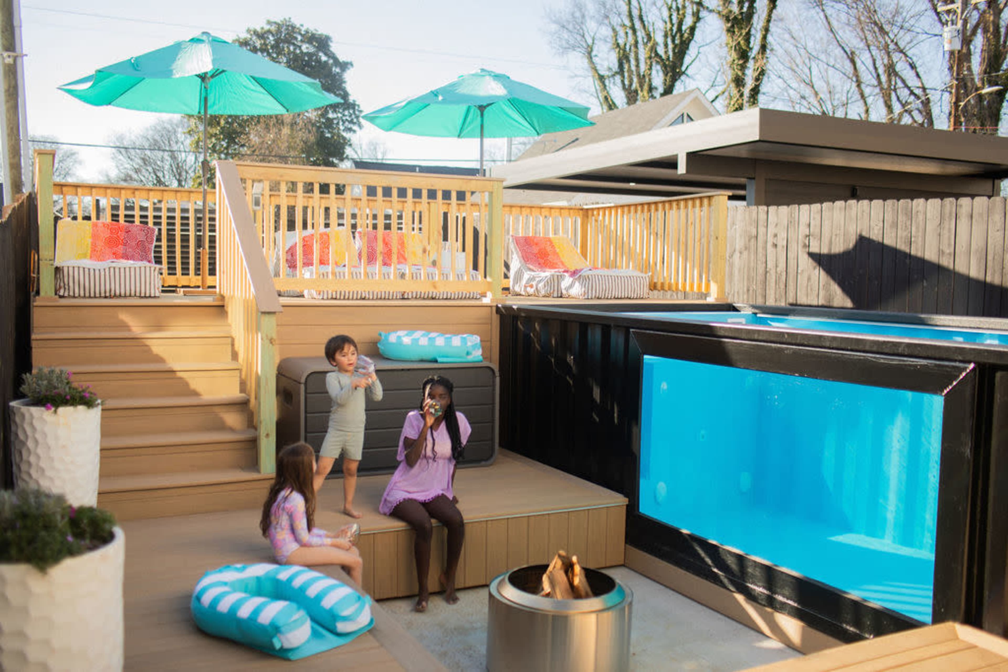 A family enjoys a sunlit backyard with a wooden deck, pool, and seating areas beneath green umbrellas.