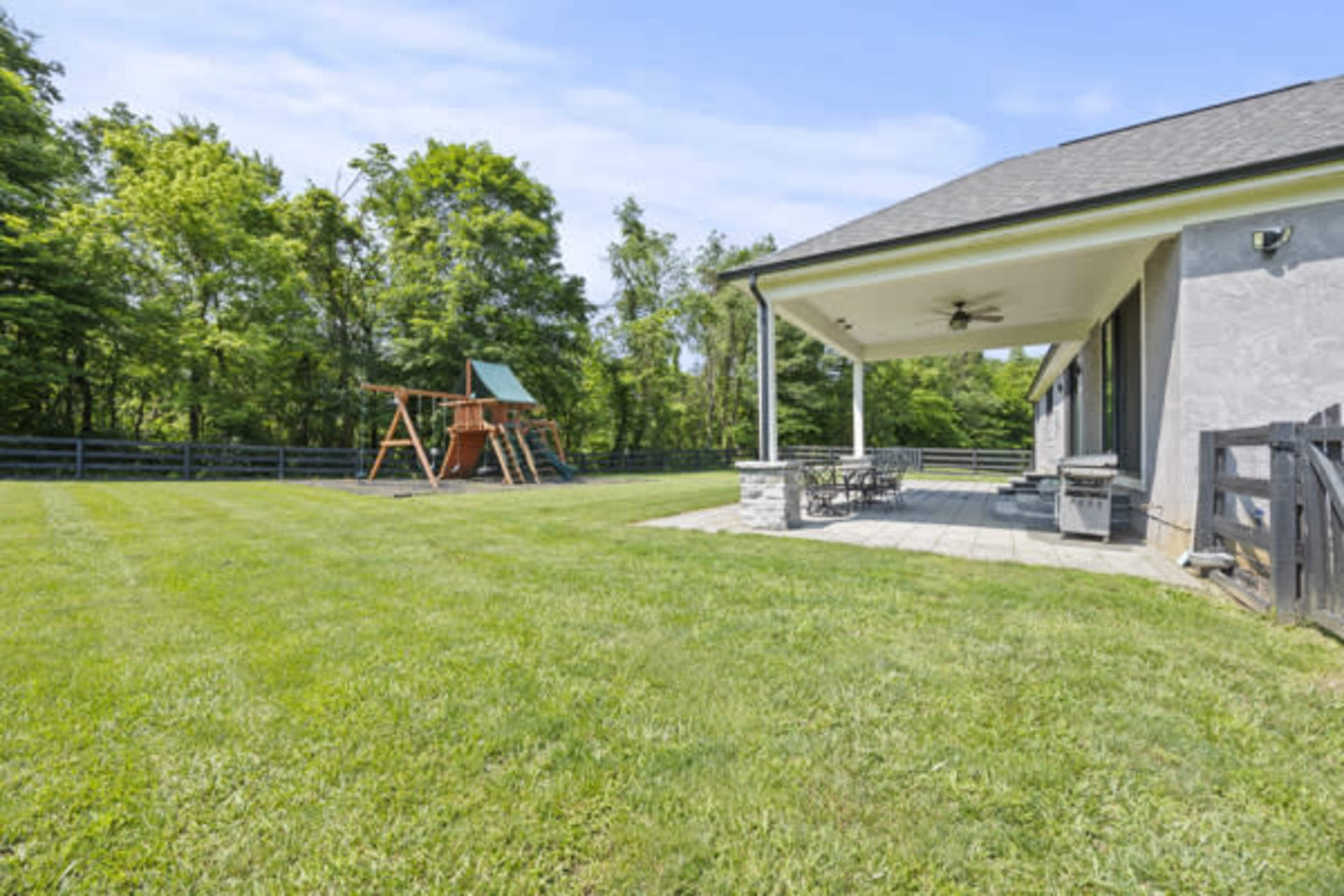 The image shows a spacious backyard featuring a wooden playset and a covered patio area with outdoor seating.