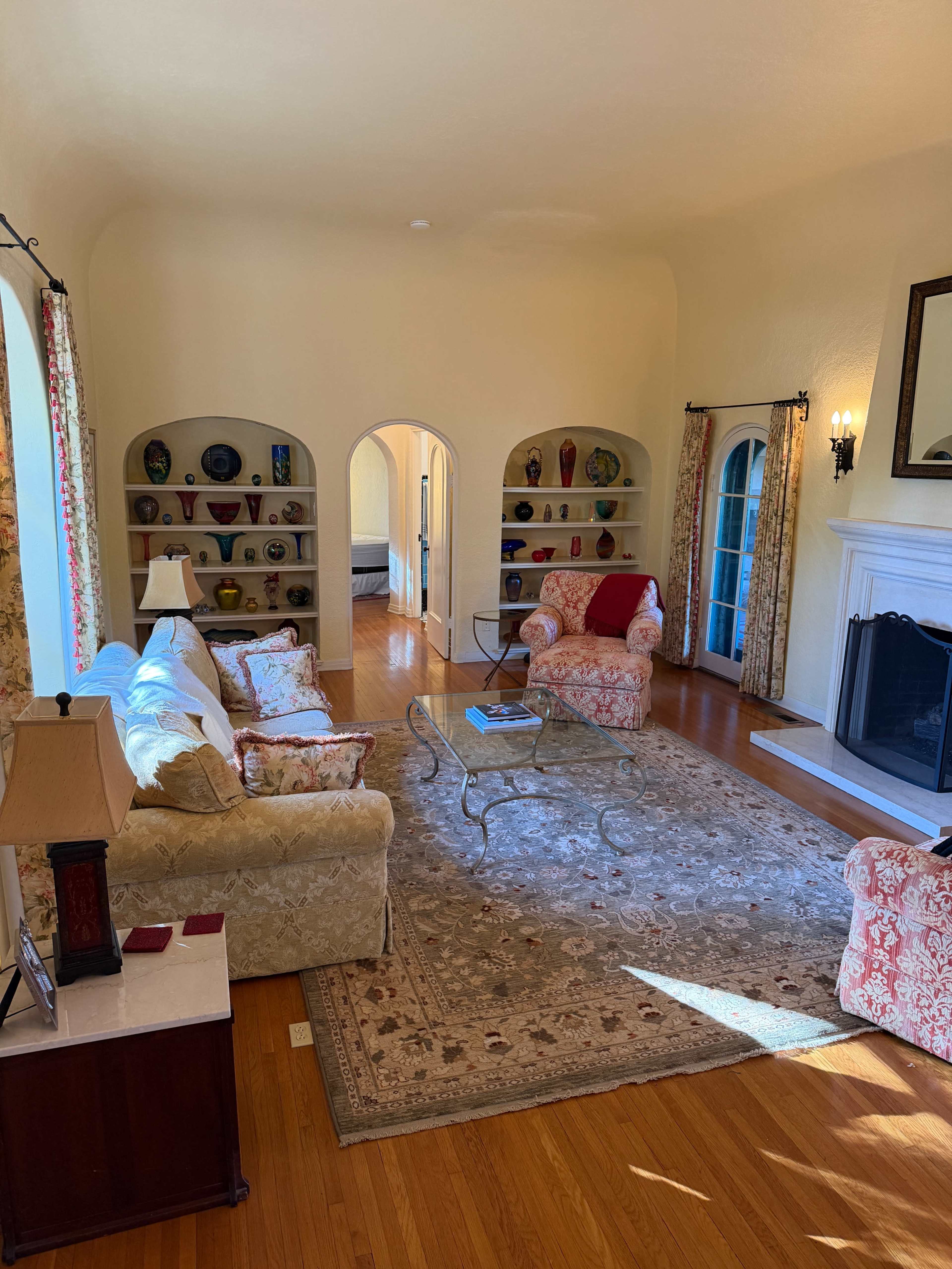 The image shows a cozy living room with a fireplace, bookshelves filled with decorative vases, and two patterned chairs arranged around a coffee table.