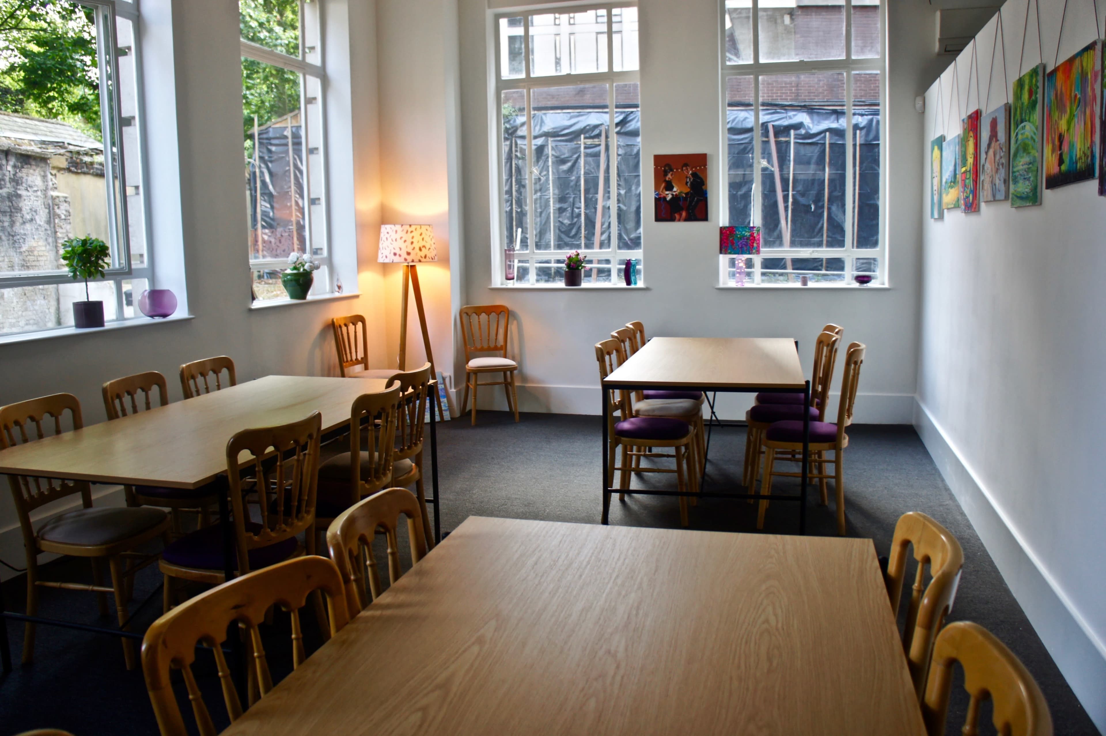 A brightly lit room features several wooden tables and chairs arranged across a carpeted floor, with colorful artwork displayed on the walls.