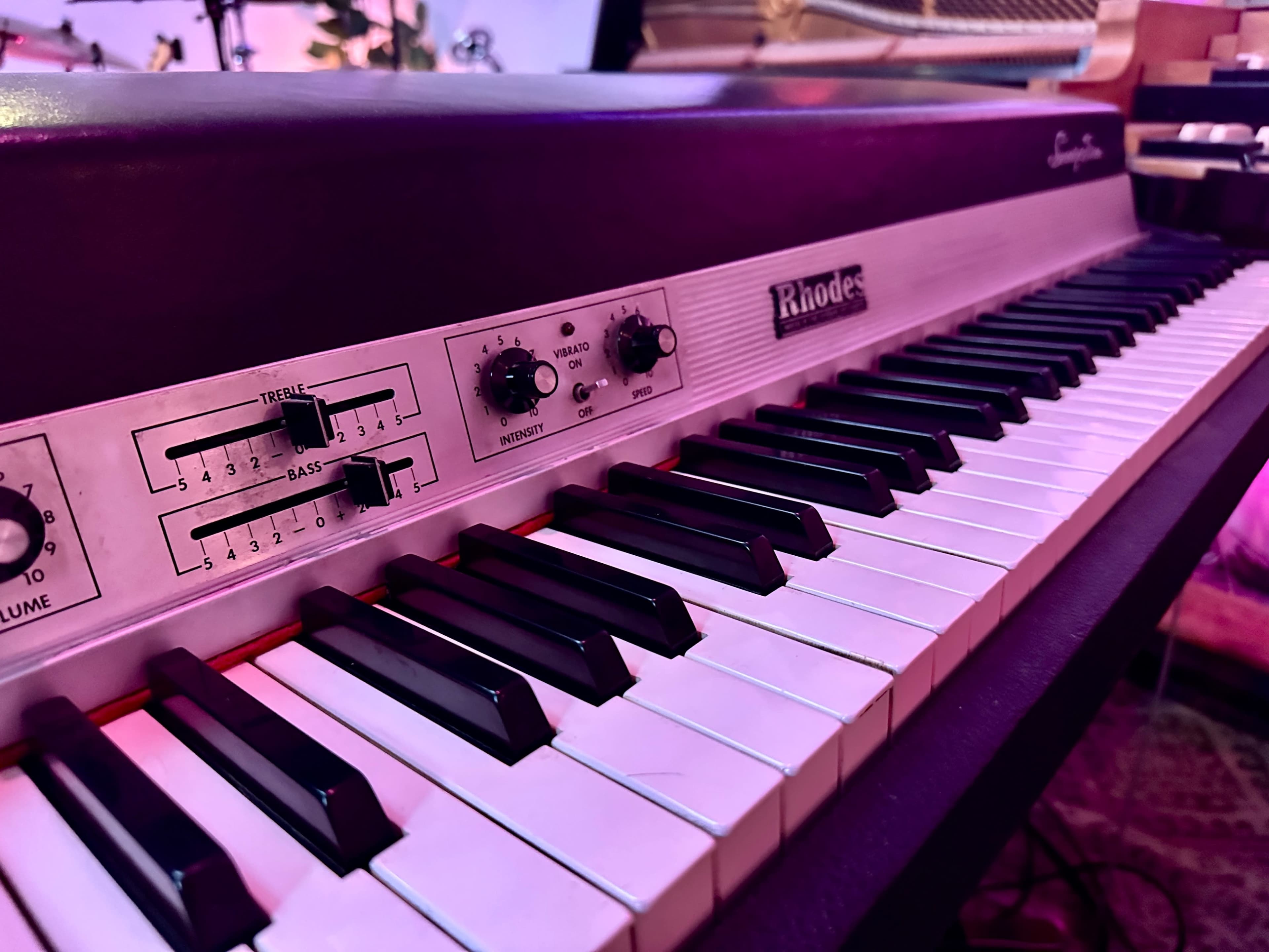 The image shows a close-up of a Rhodes electric piano with black and white keys and control knobs visible on the left side.