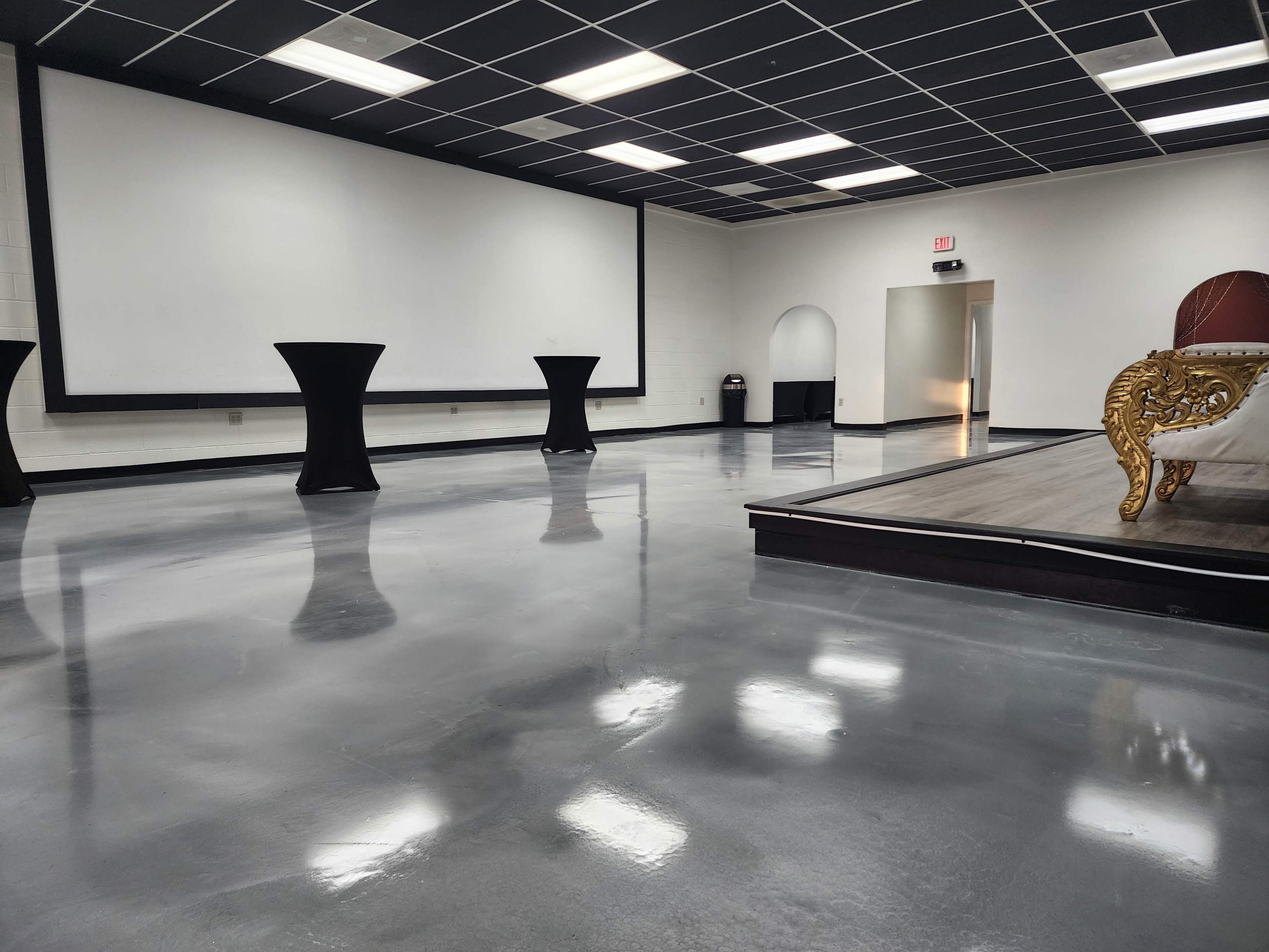 A spacious event room features polished concrete floors, a large projection screen, and several round black tables.