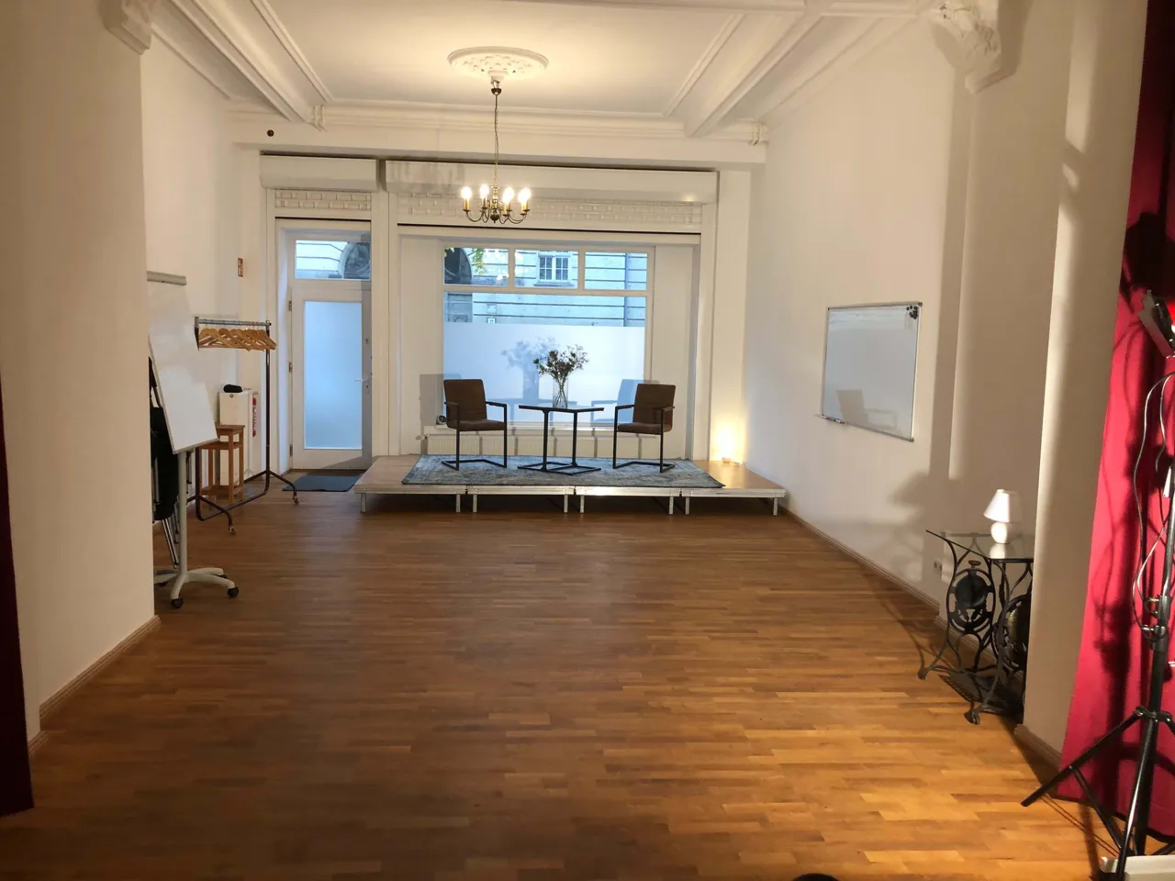 The image shows a spacious, well-lit room with wooden floors, featuring a stage area with two chairs and a table, flanked by a whiteboard and decorative lamps.