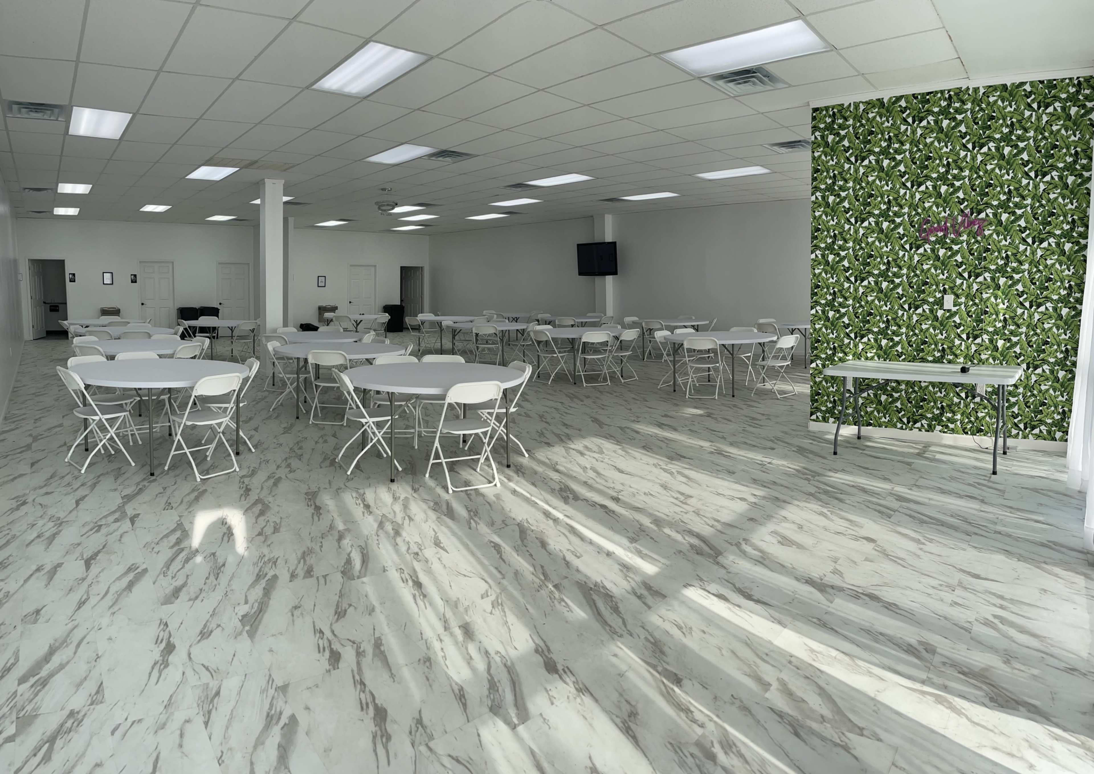 A spacious event room with multiple round tables and chairs arranged neatly, featuring a decorative green wall on one side and sunlight casting shadows on the marble floor.