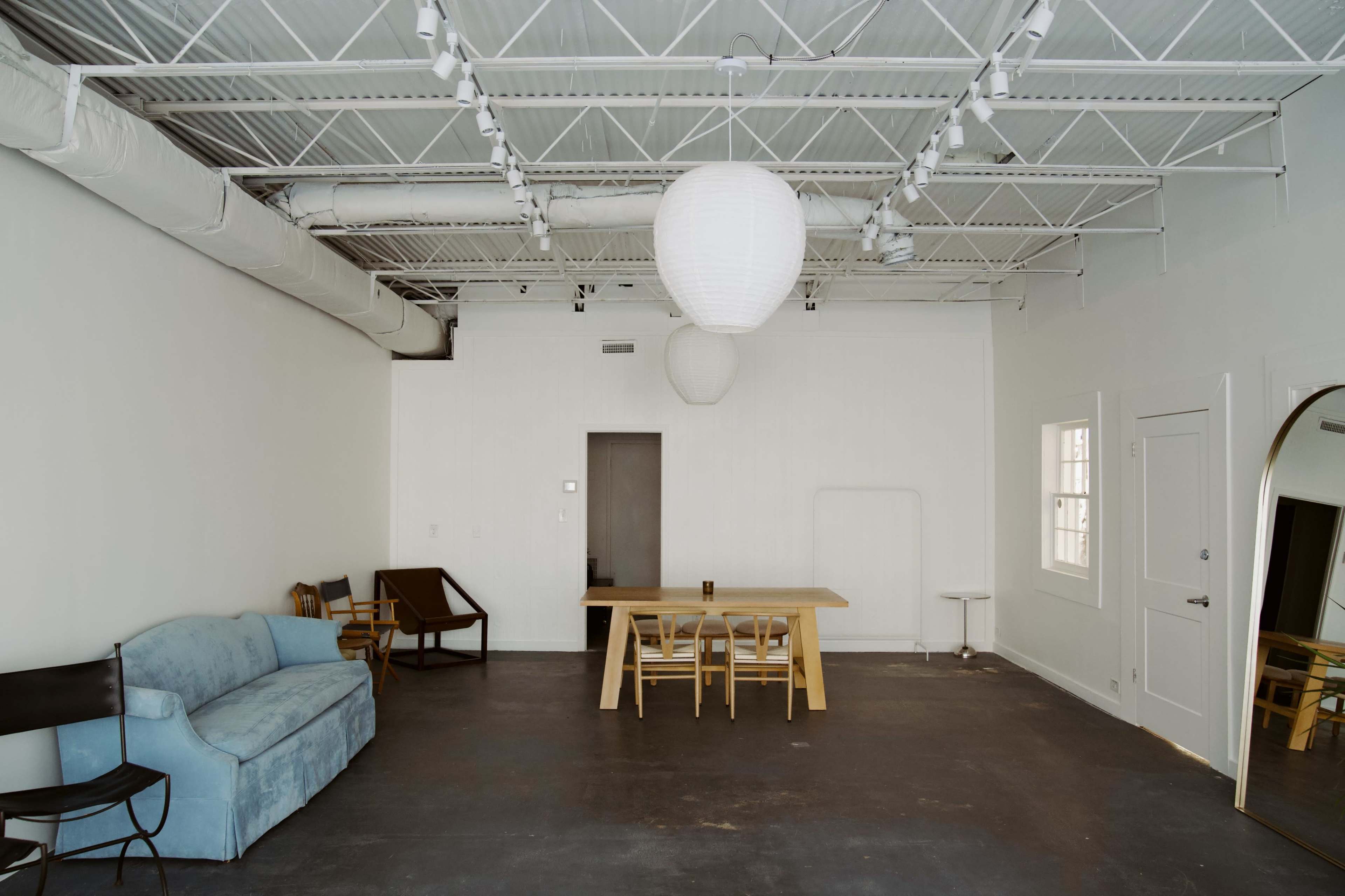 The image shows a minimalist interior space with a blue couch, a wooden dining table surrounded by chairs, and two paper lanterns hanging from the ceiling.