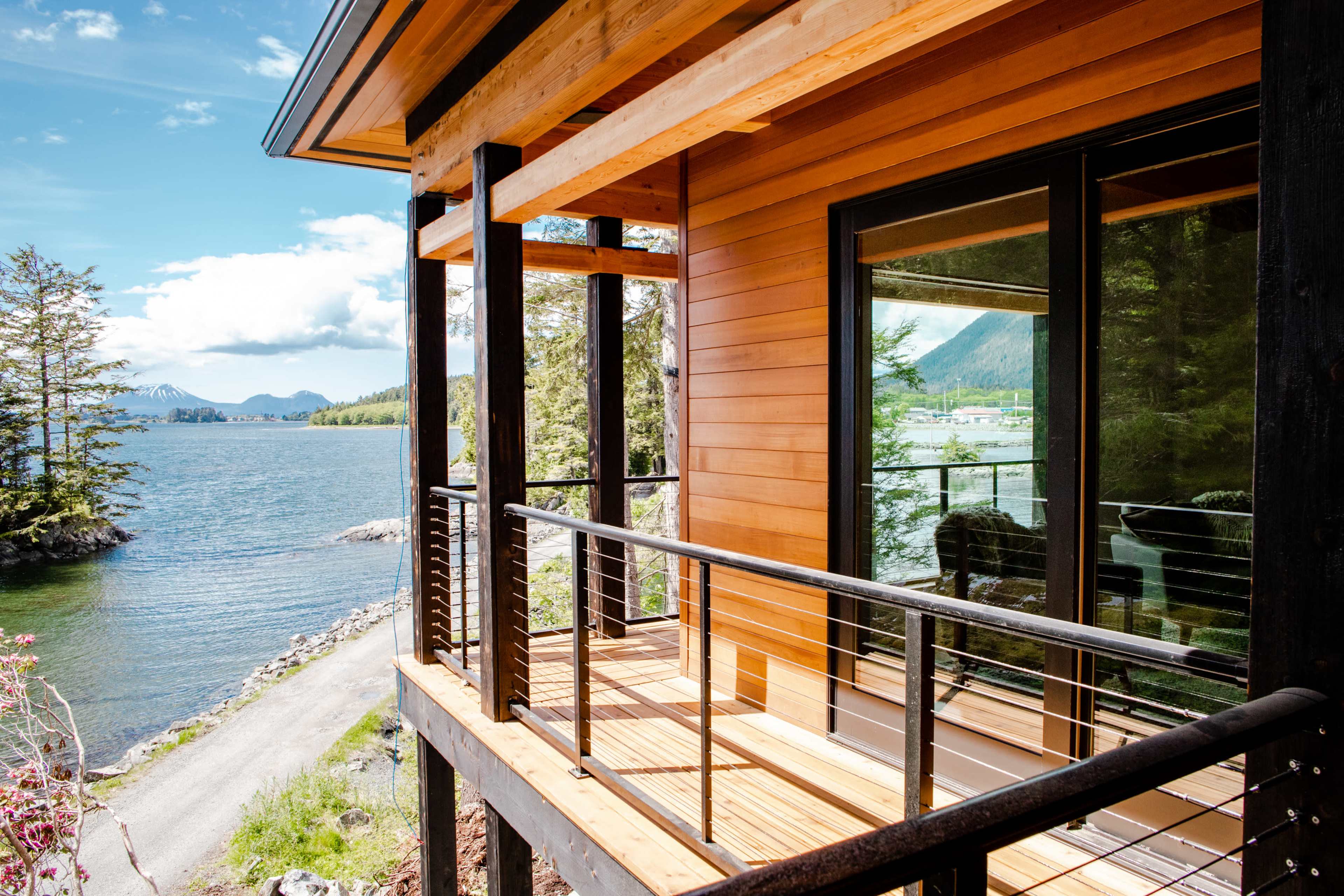 A wooden balcony overlooks a calm body of water surrounded by trees and distant mountains.