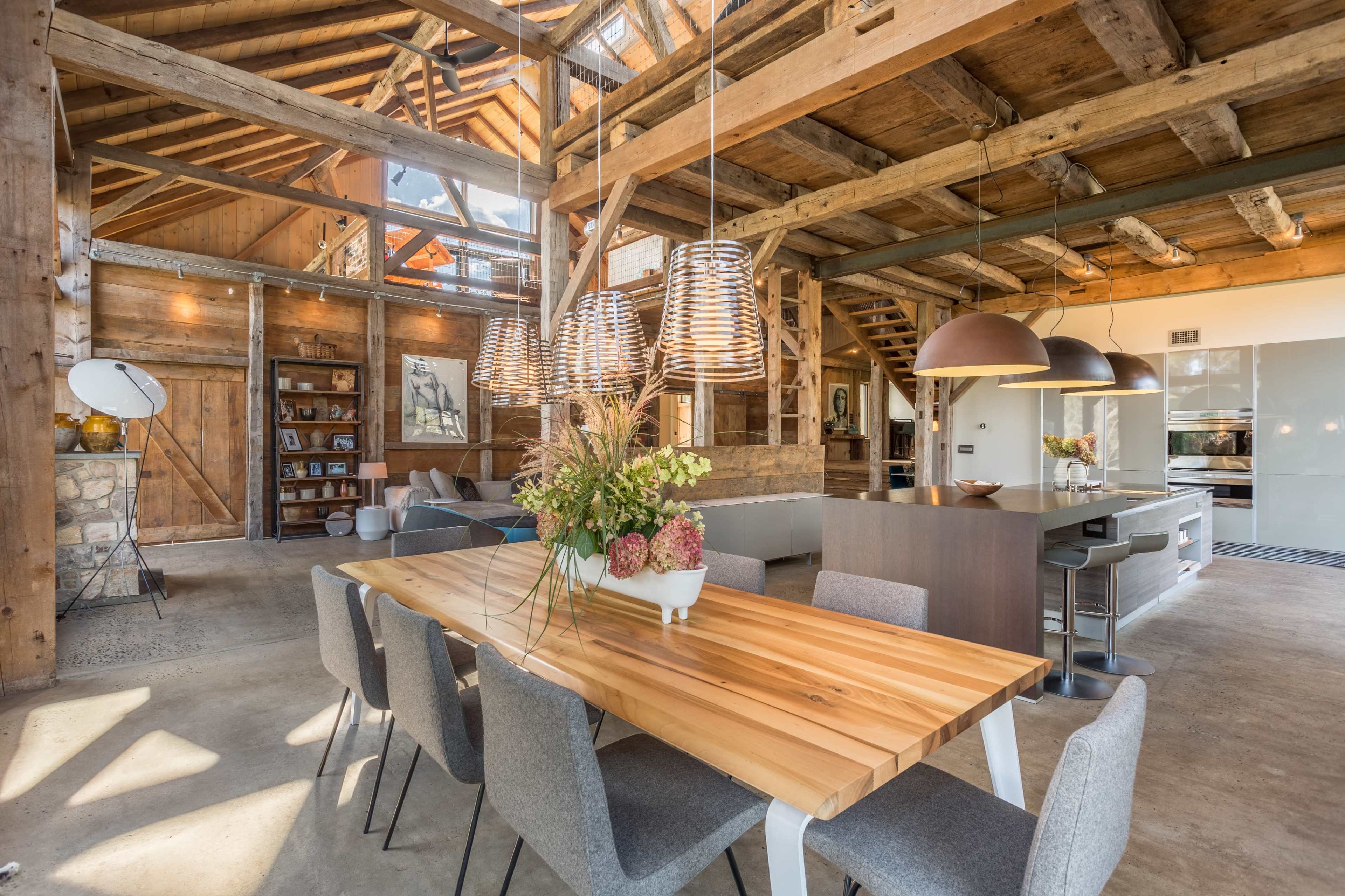 A spacious, rustic interior with wooden beams, a large dining table surrounded by chairs, and an open kitchen area.
