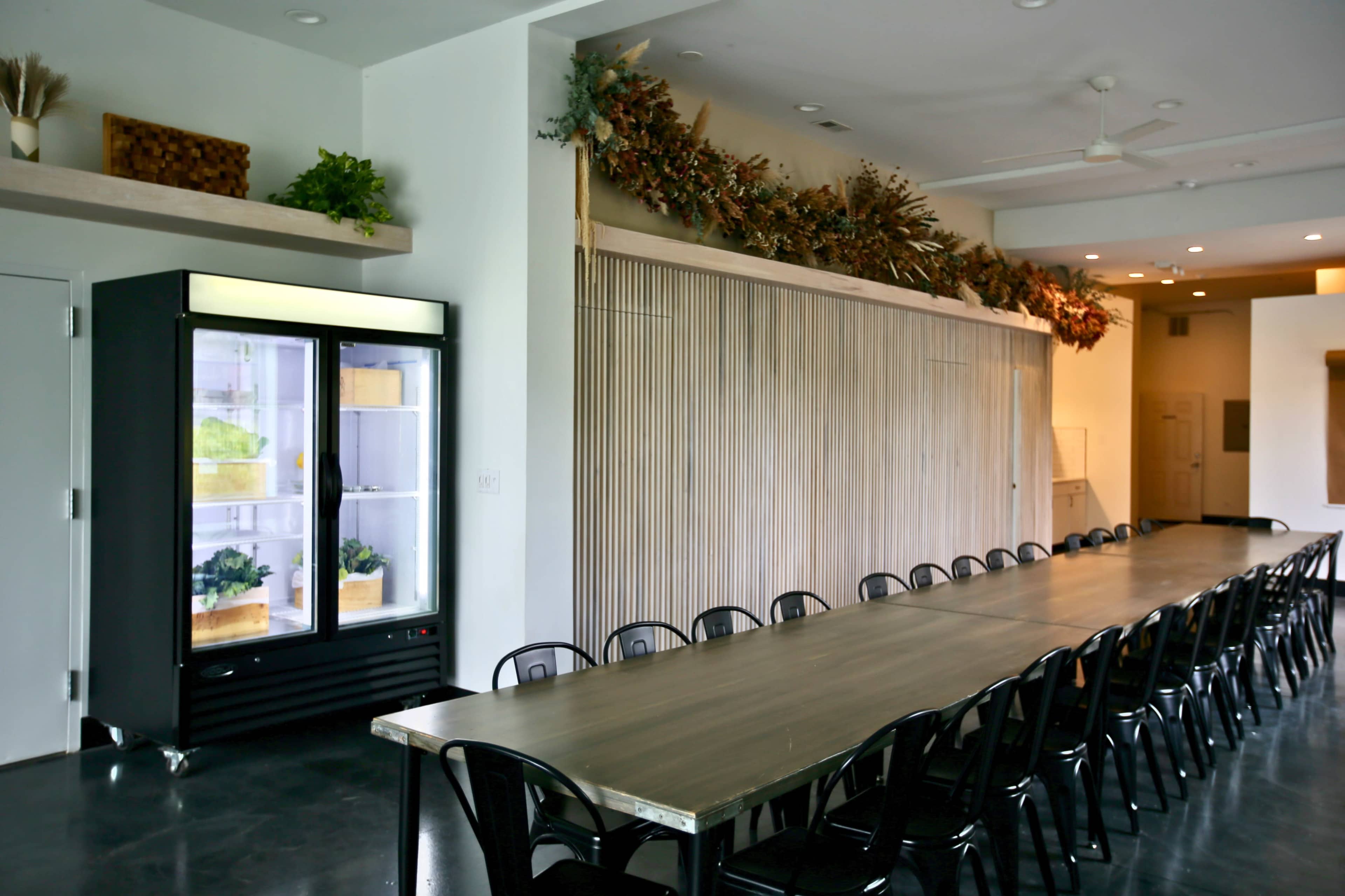 A spacious room with a long wooden table surrounded by black chairs, a refrigerator in the corner, and decorative plant arrangements on the walls.