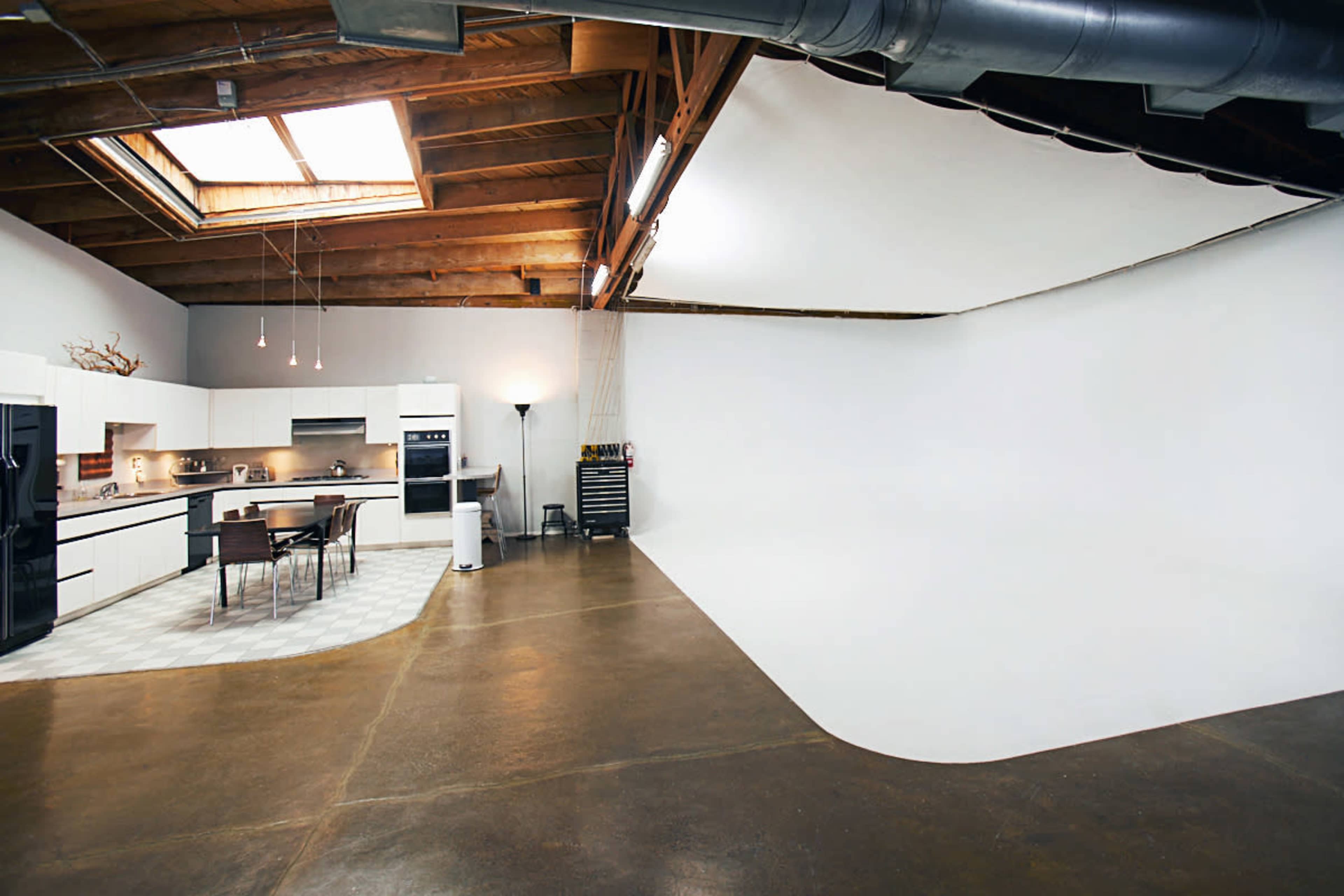 A spacious kitchen with modern appliances, a dining table, and a large white backdrop in an open area with wooden beams and a skylight.