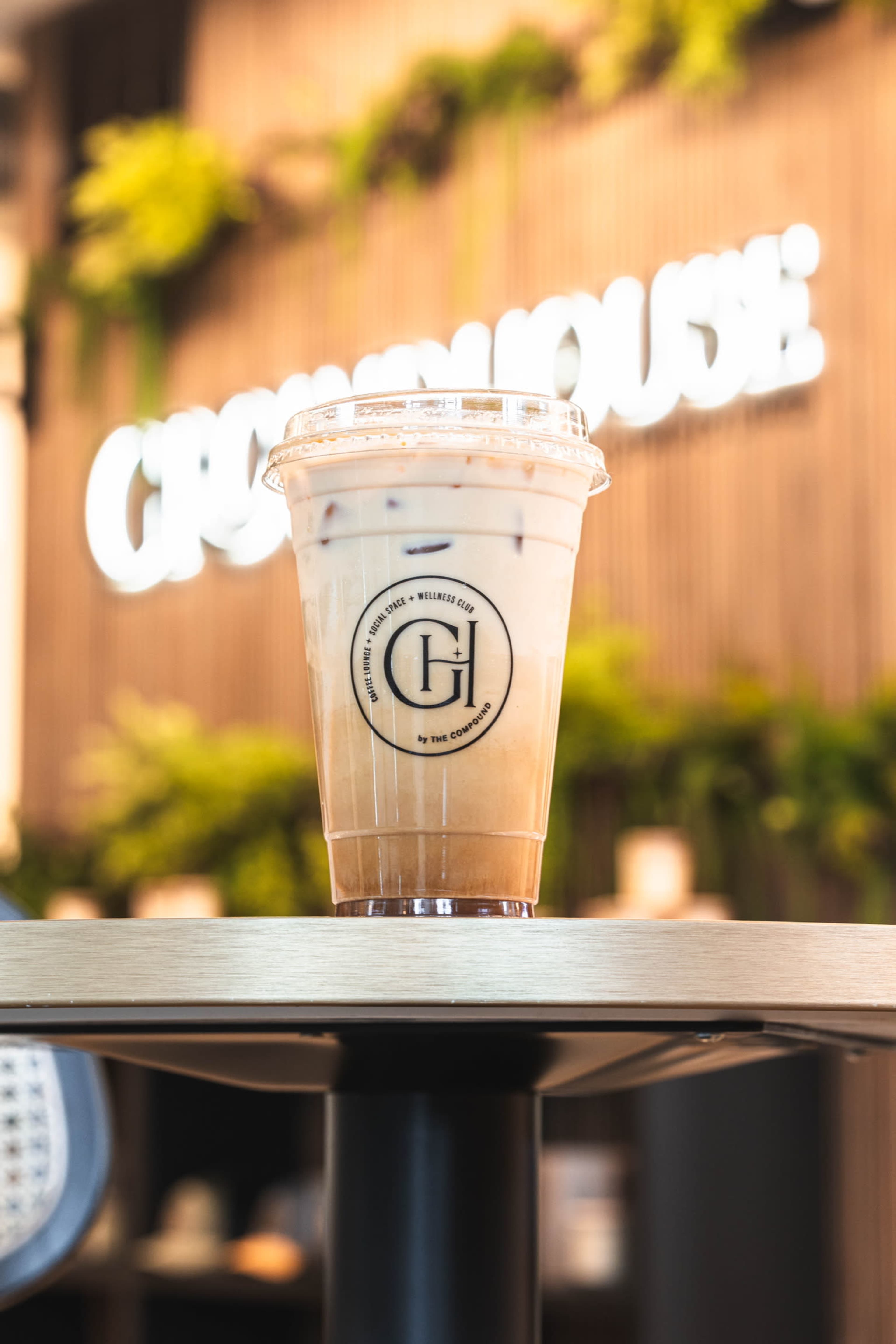 A clear plastic cup filled with iced coffee sits on a table in front of a blurred background featuring greenery and a sign that reads "GREENHOUSE."