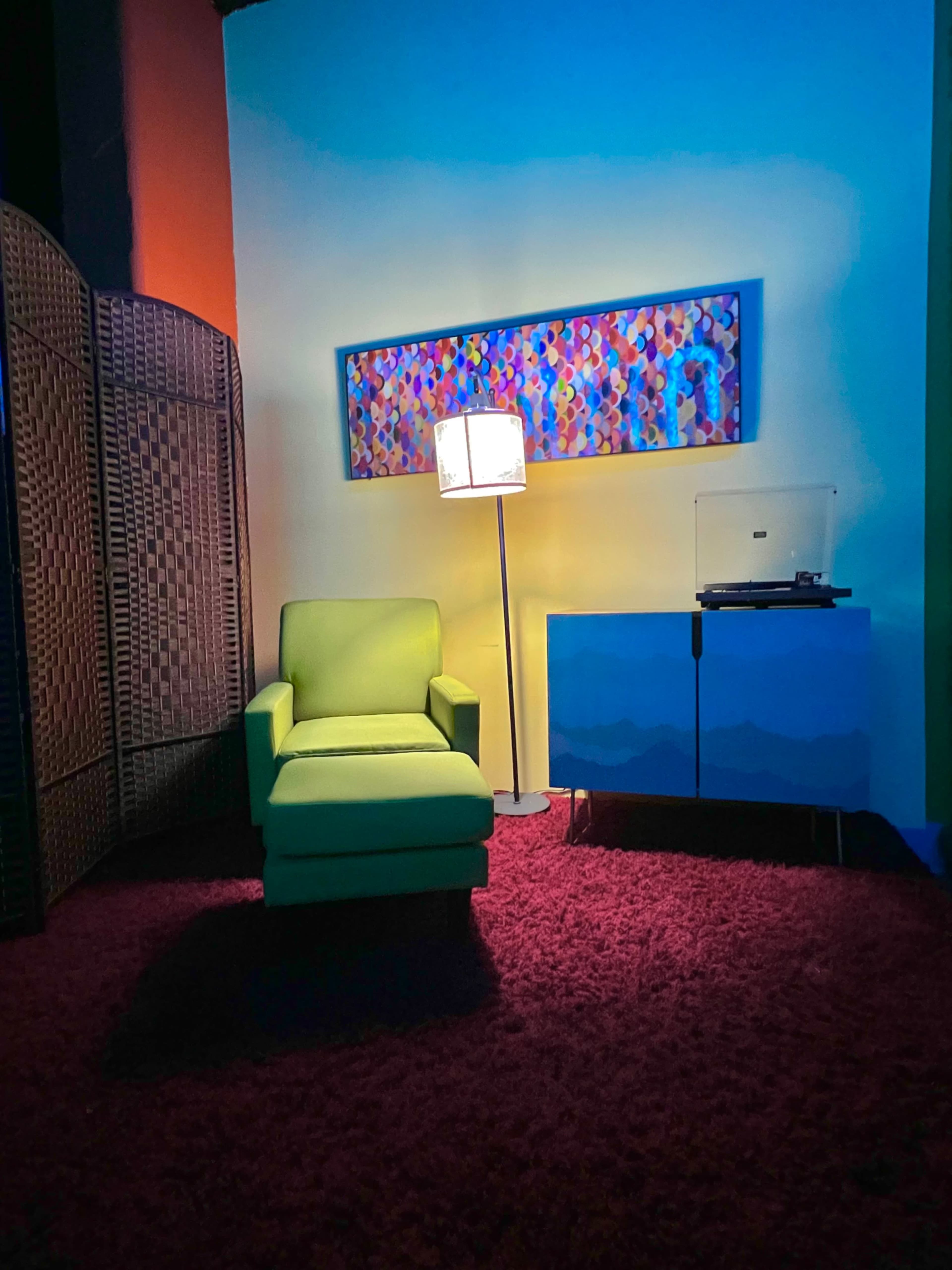 A green armchair is positioned next to a lamp and a turntable, set against a colorful wall art piece and a woven screen in a room with a red carpet.