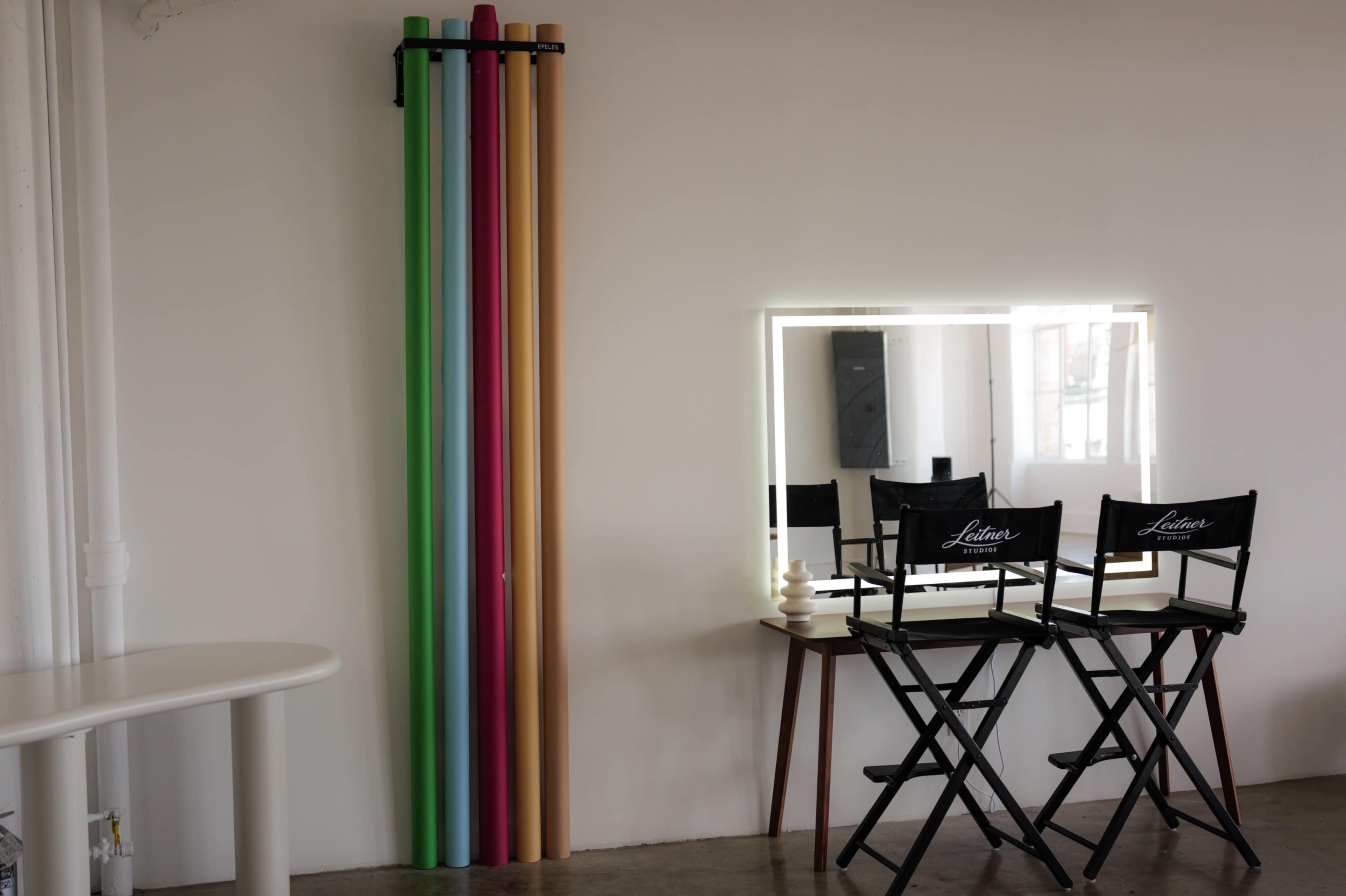 The image shows a minimalistic interior featuring a set of colored backdrop rolls mounted on the wall next to a vanity mirror and two director’s chairs.