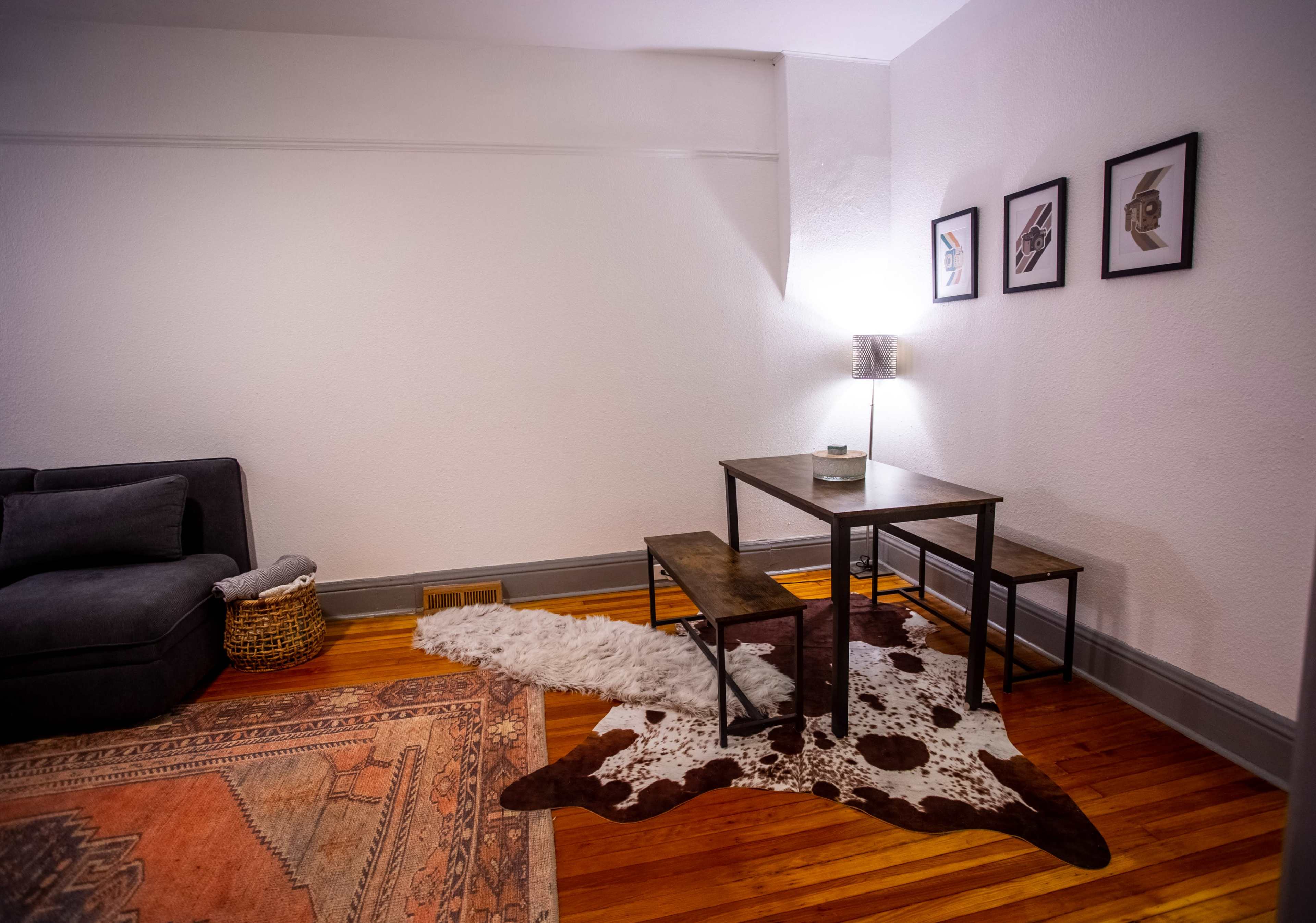 A living space with a gray couch, a small dining table with benches, a lamp, and a cowhide rug on a wooden floor.
