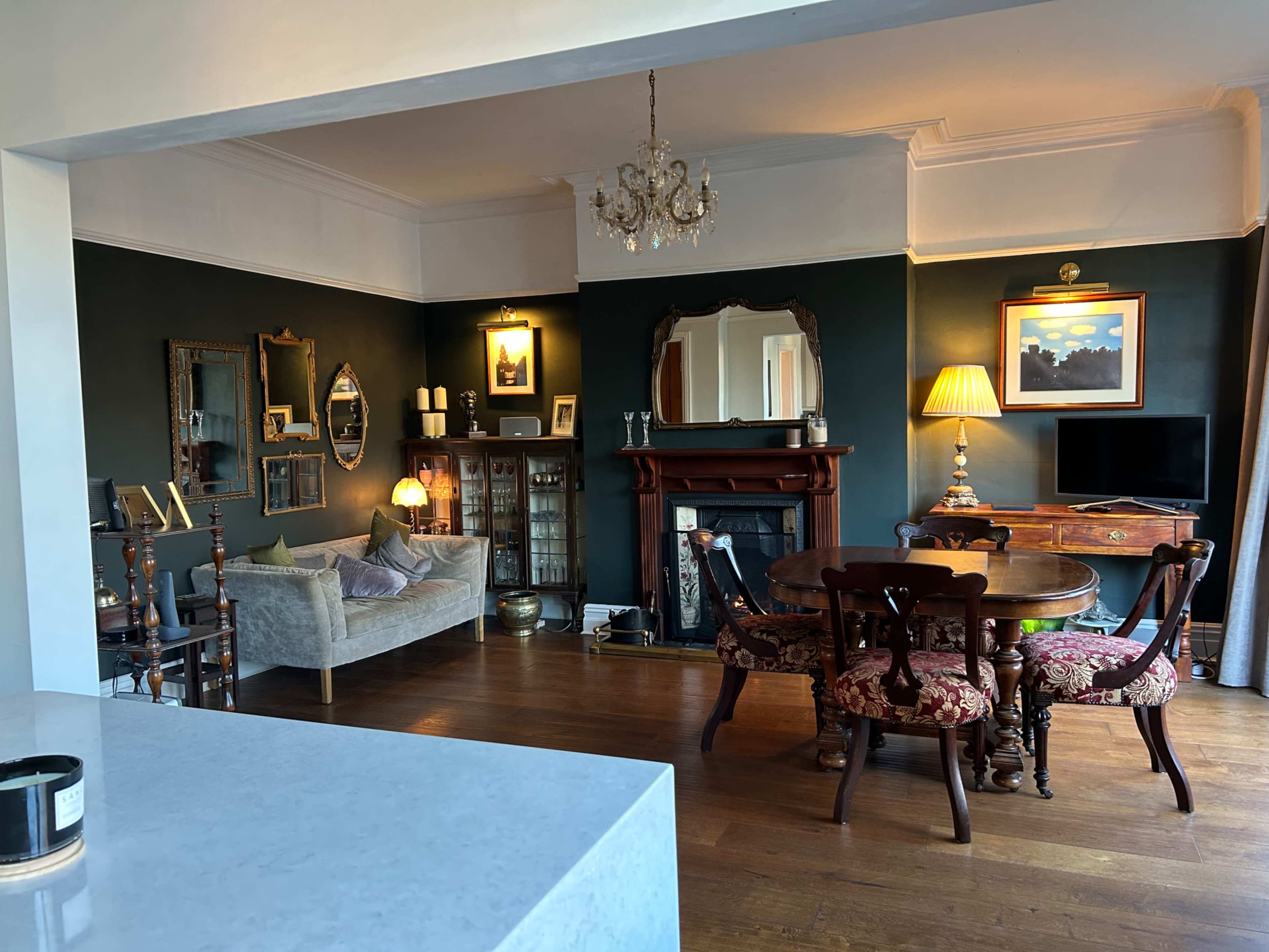 The image shows a cozy living room with a mix of antique furniture, including a sofa, dining table, and a decorative fireplace.