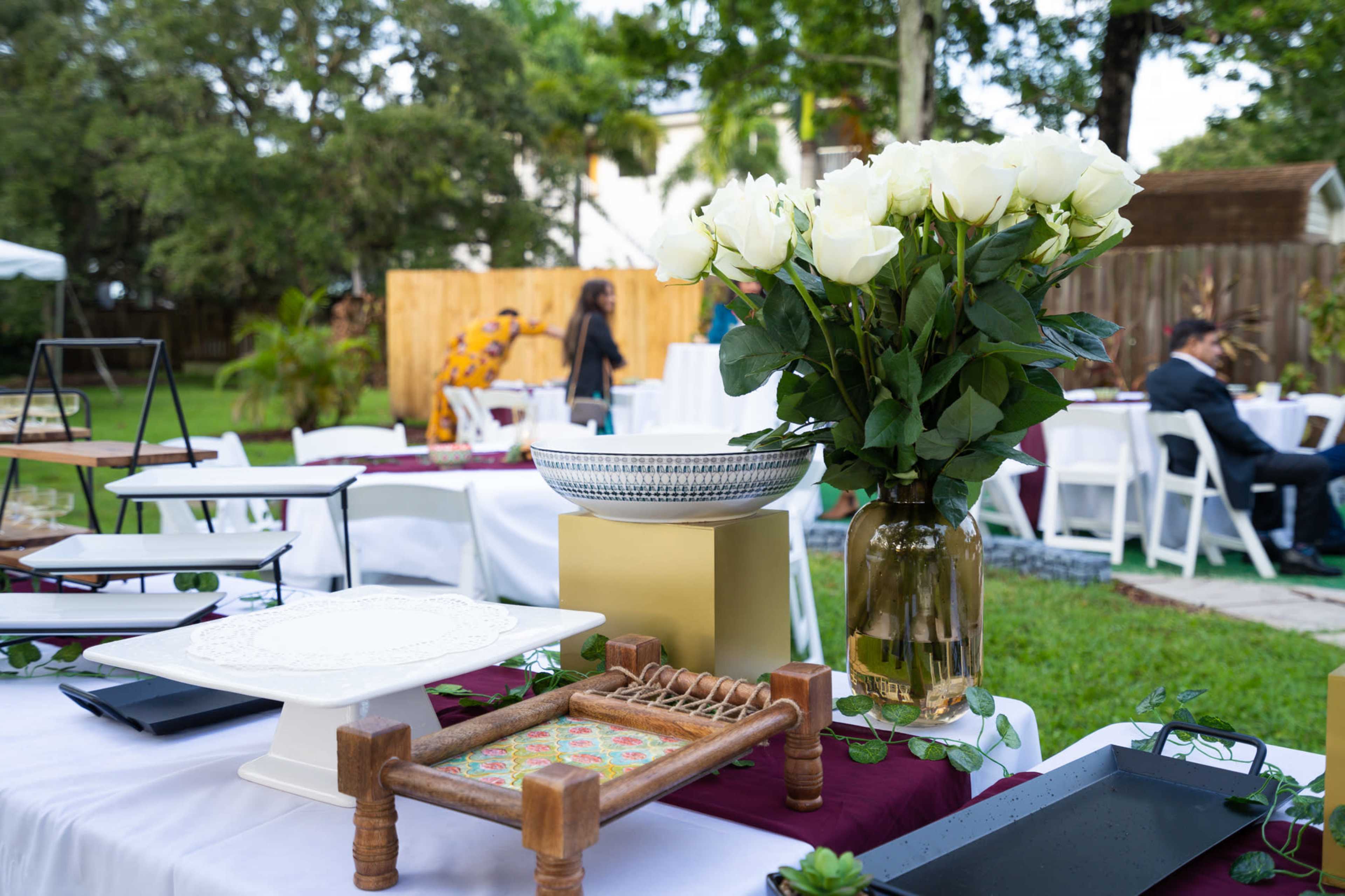 A decorated outdoor setting with white tables, floral arrangements, and various serving trays arranged for an event.
