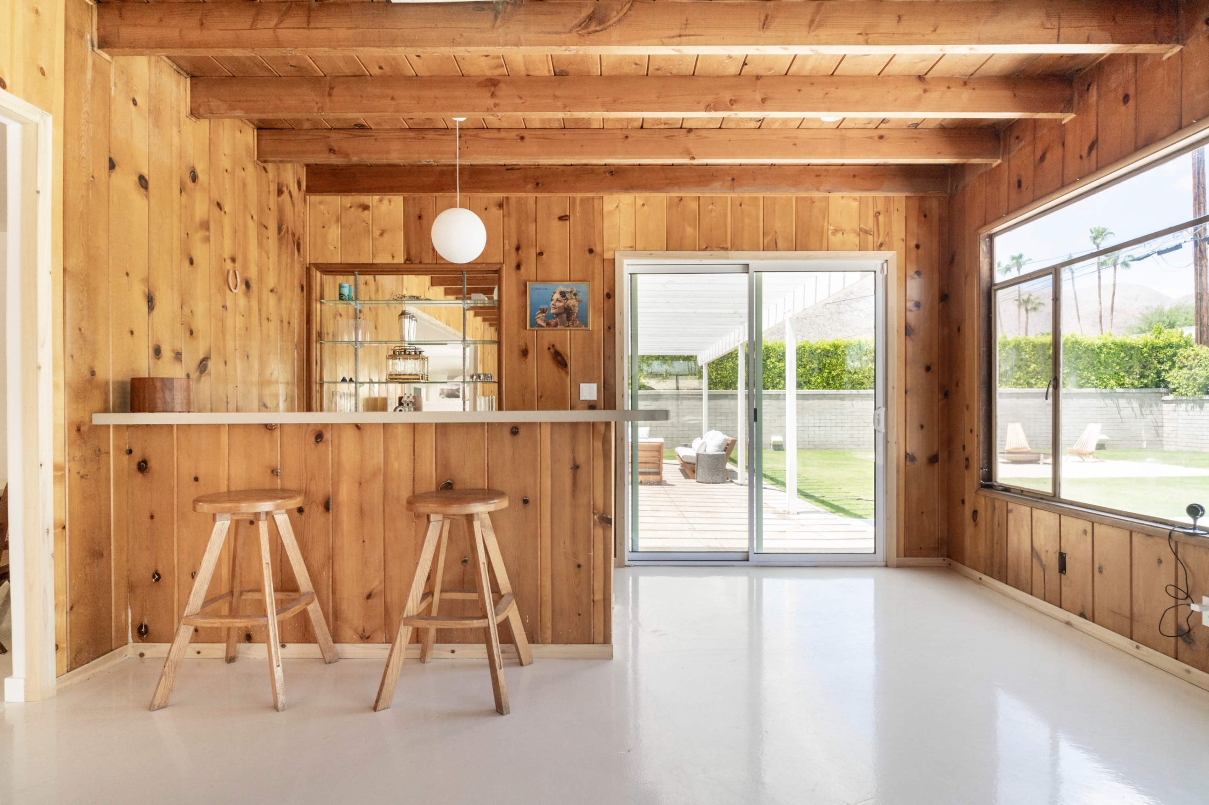 A wooden-paneled interior featuring a bar with two stools, a glass sliding door, and a view of an outdoor patio area.