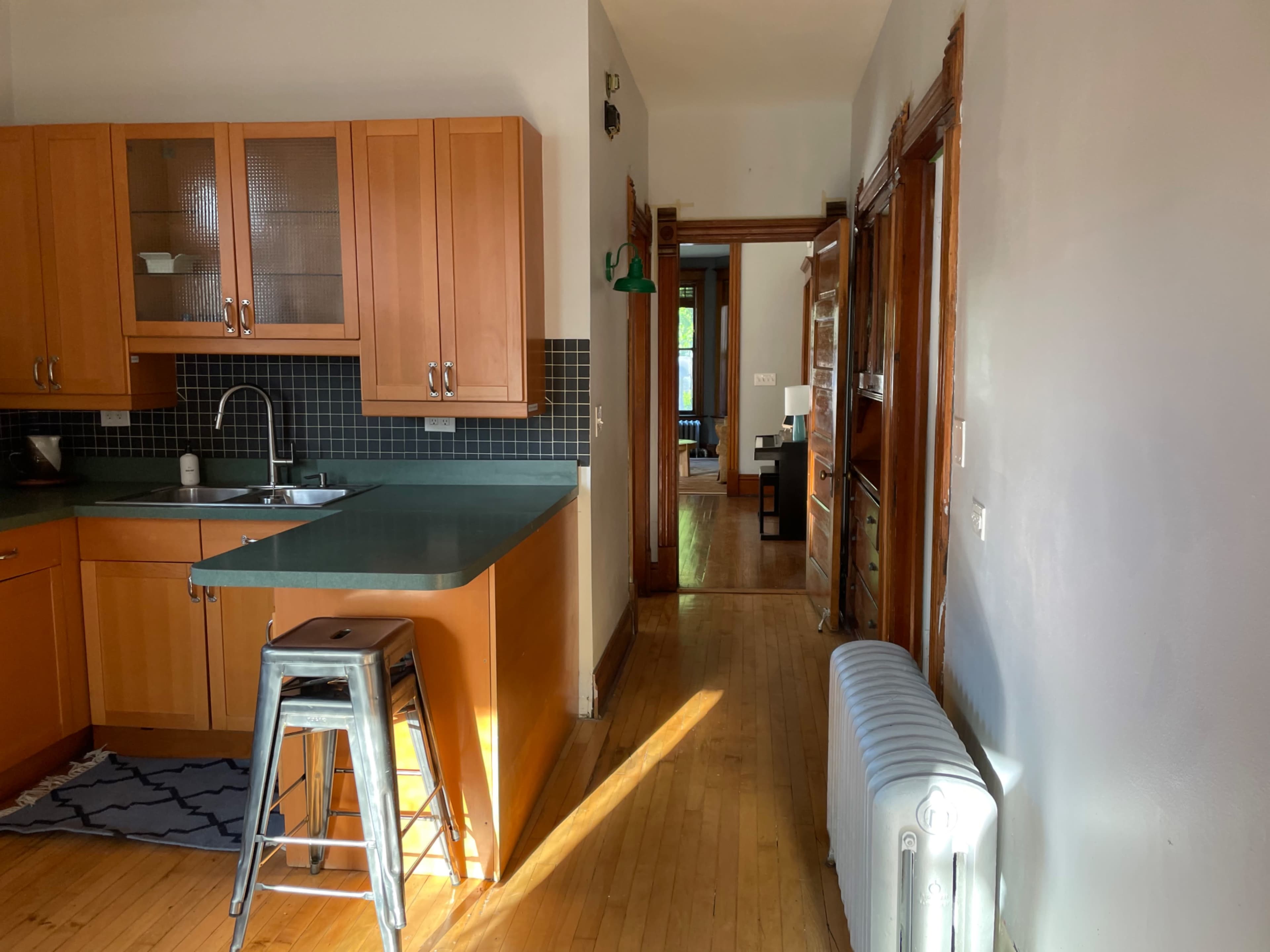 A bright kitchen with wooden cabinets, a green countertop, and a hallway leading to a living area is visible.