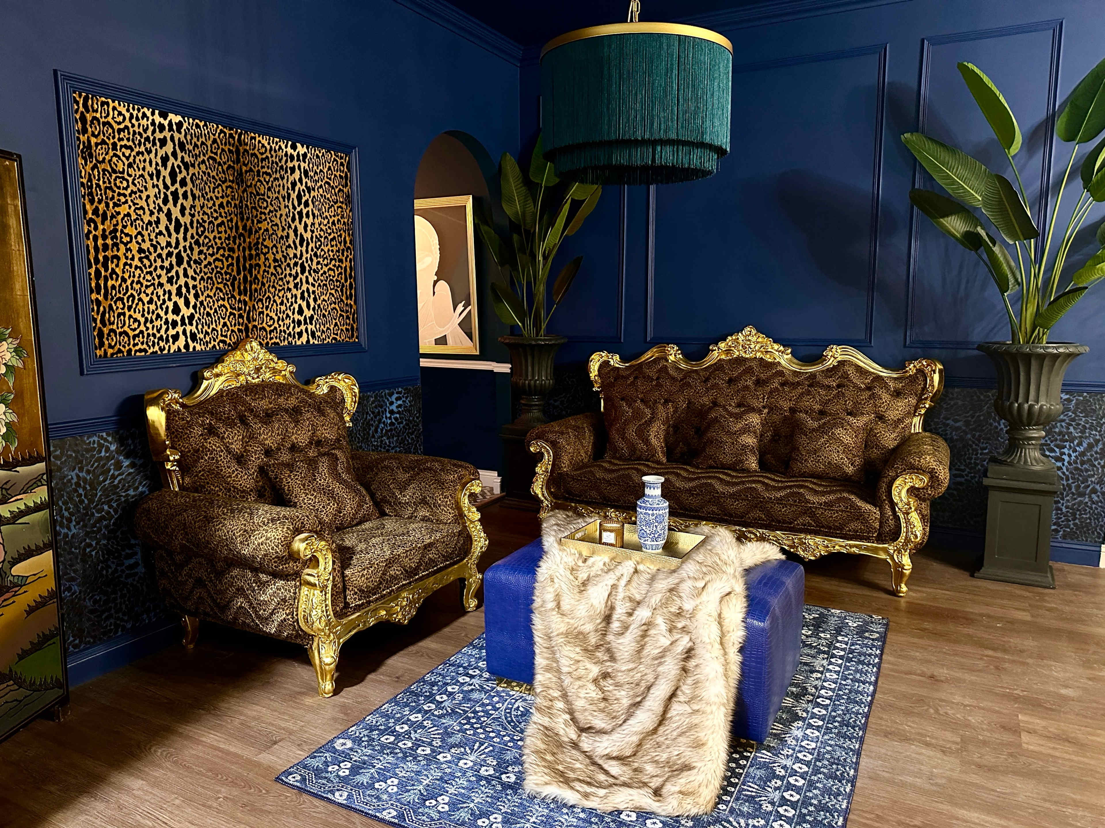A luxurious living room with ornate gold-framed sofas, a blue coffee table, and tall green plants against a dark blue wall.