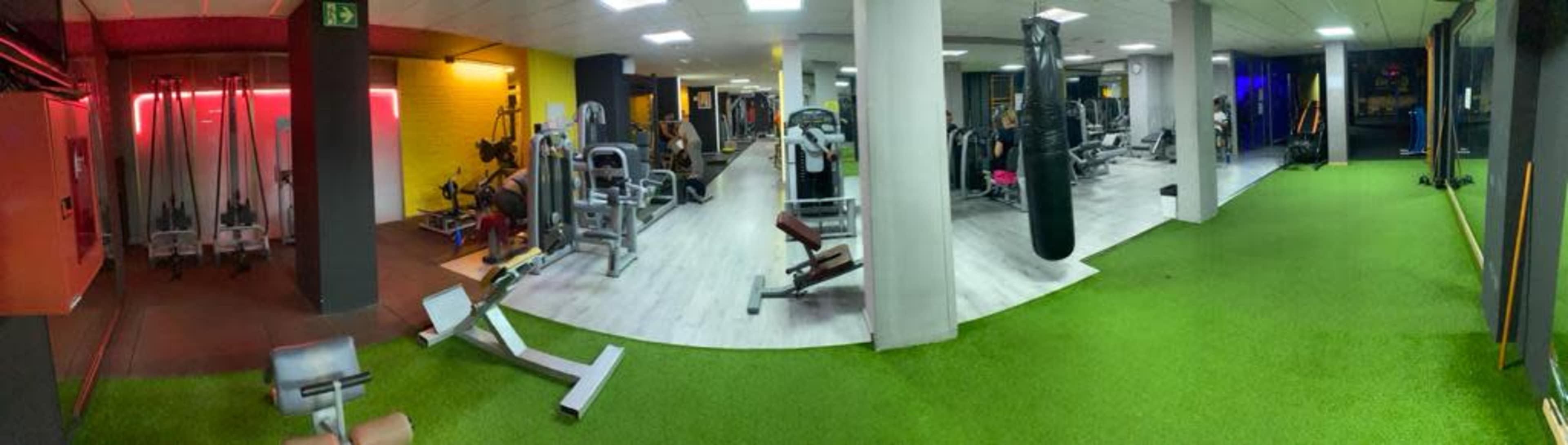 A panoramic view of a gym interior featuring various exercise equipment, including weight machines, a boxing bag, and workout spaces with artificial turf.