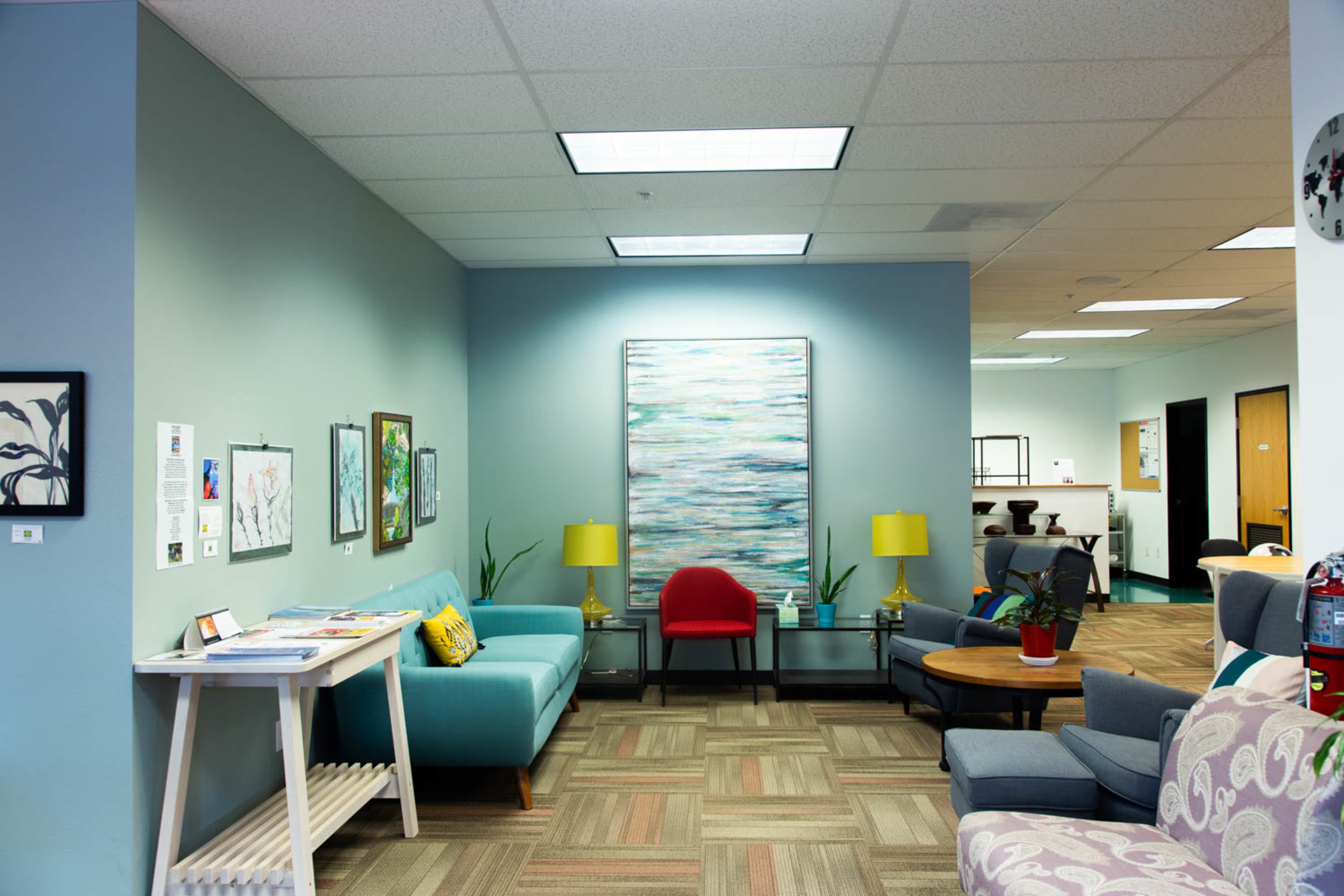 A modern waiting room with a light blue wall, a teal sofa, a red chair, and various artworks, illuminated by ceiling lights.
