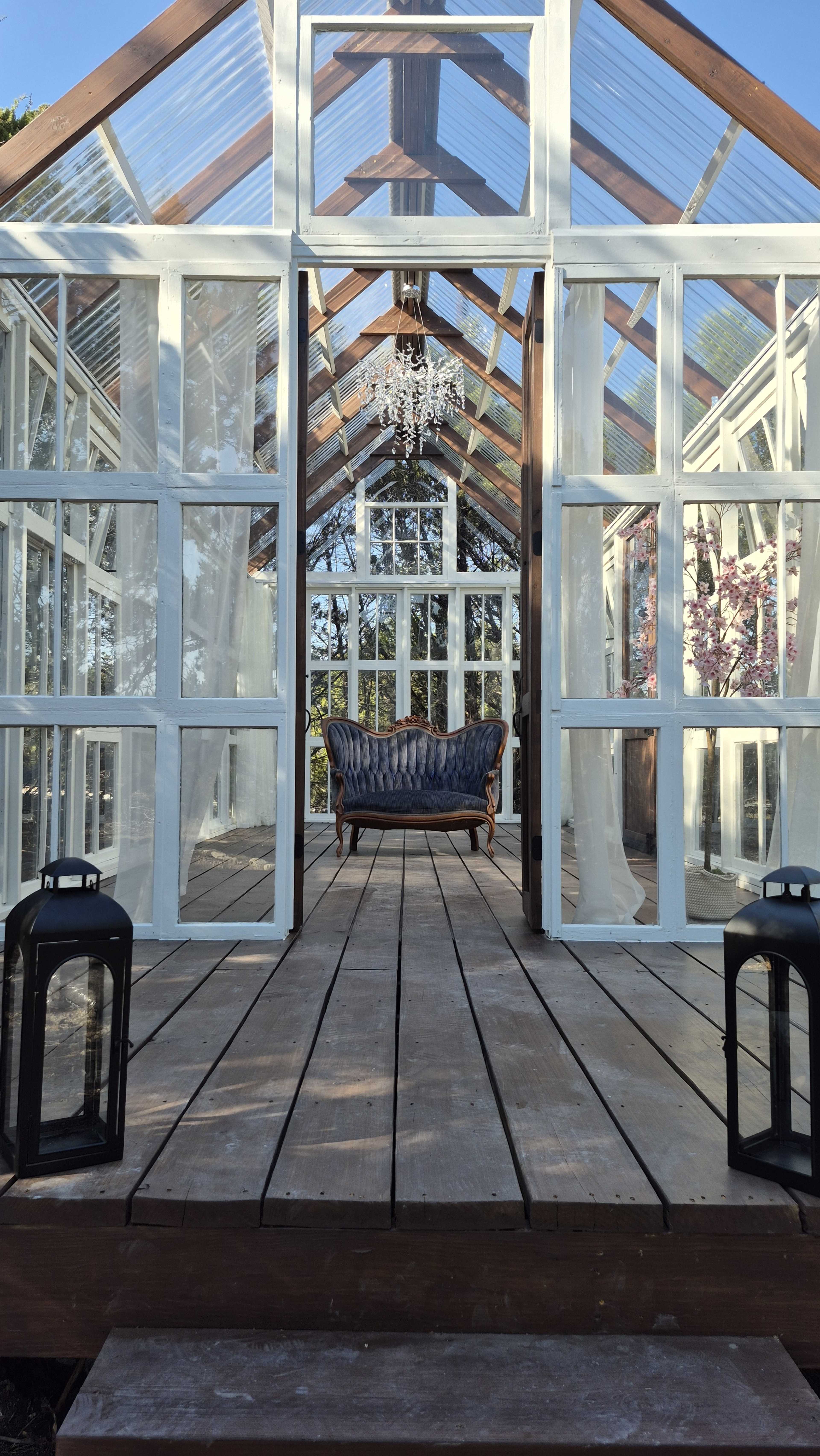 A glass greenhouse features wooden flooring, lanterns on each side, and a vintage sofa positioned beneath a chandelier.