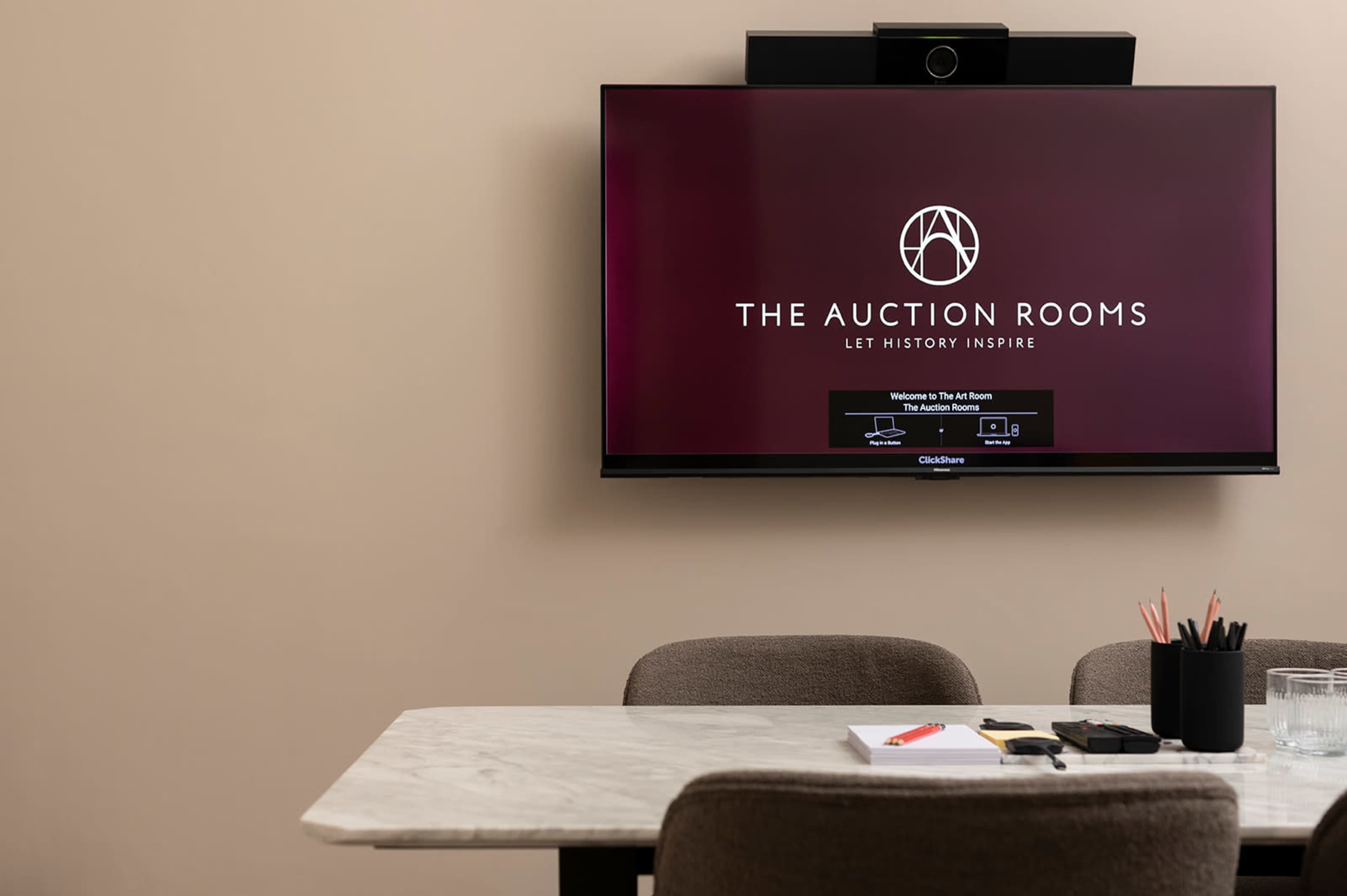 A modern conference room features a large television displaying the logo of "The Auction Rooms" above a marble table with stationery and a container of pencils.