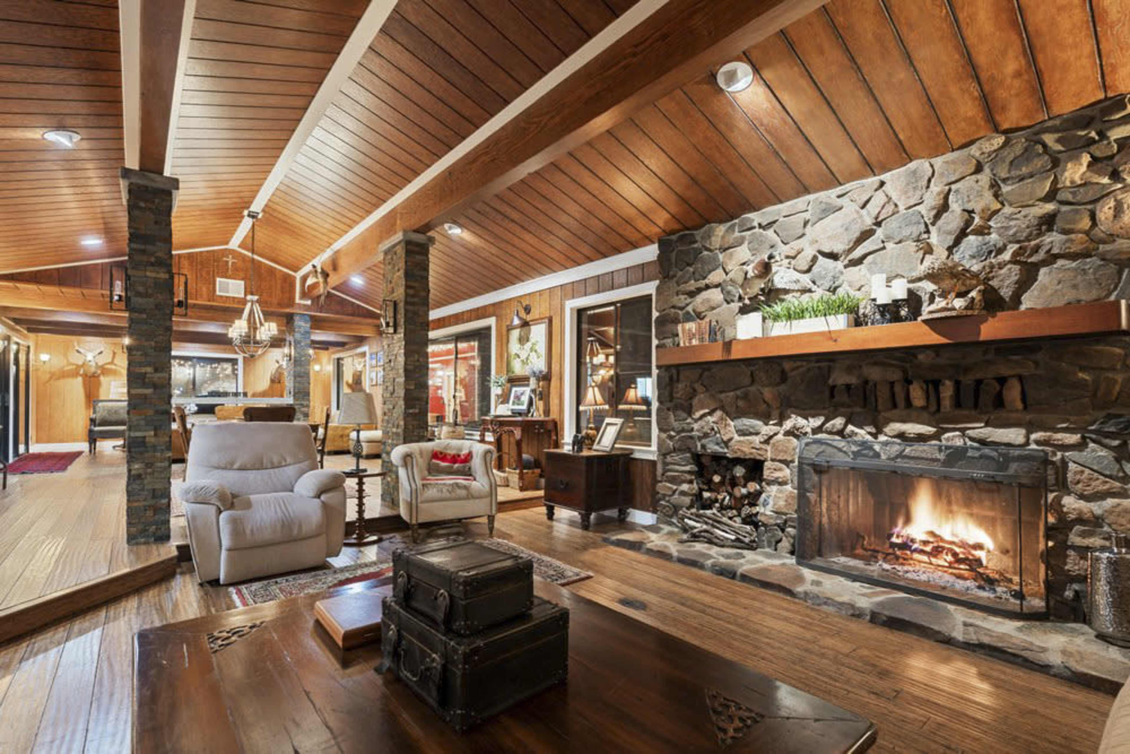 The image shows a cozy living room with a stone fireplace, wooden ceiling beams, and comfortable furniture.