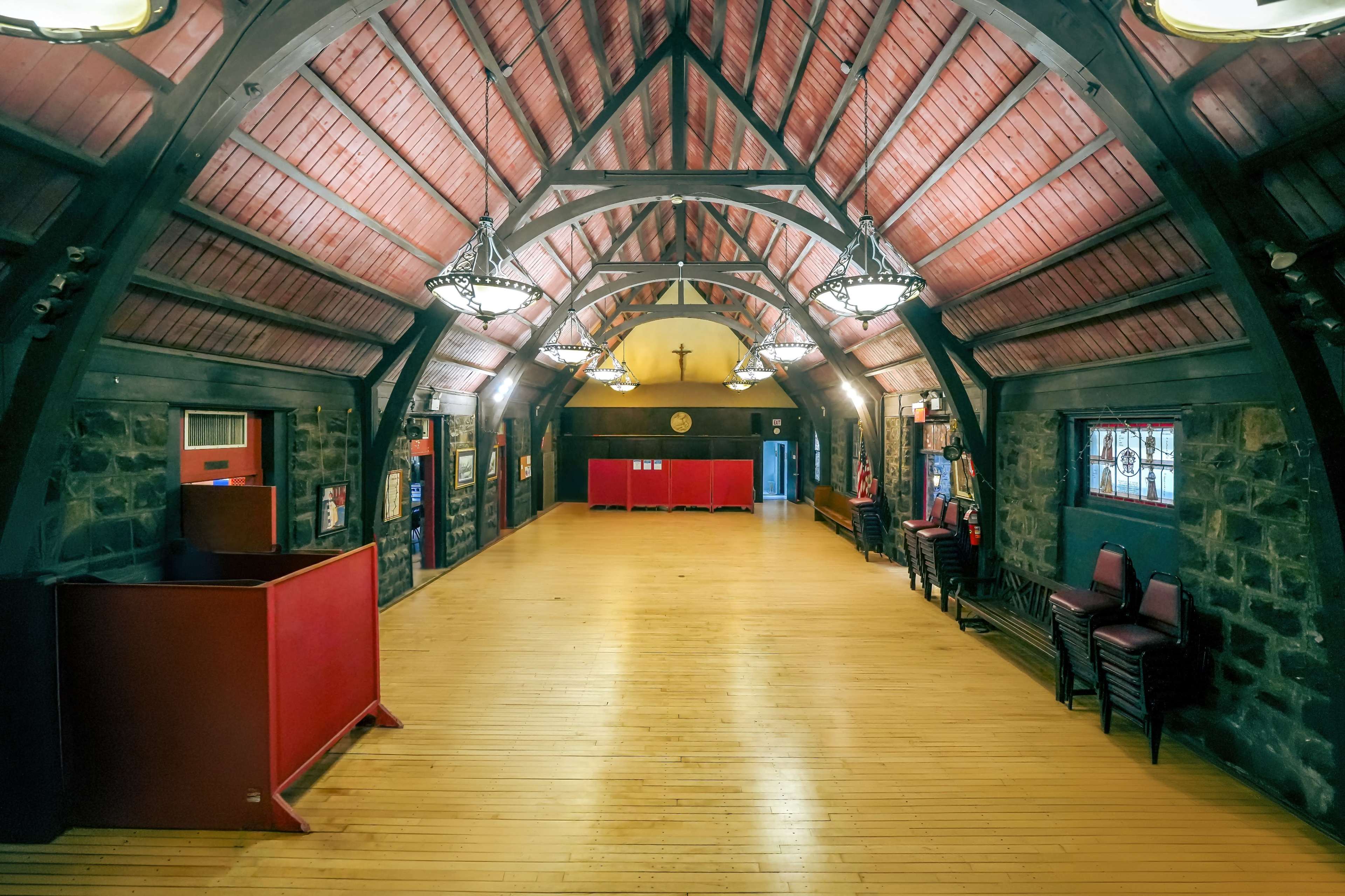 A large, open hall with a wooden floor, vaulted red wooden ceiling, and stone walls, featuring several light fixtures and red dividers along the back wall.