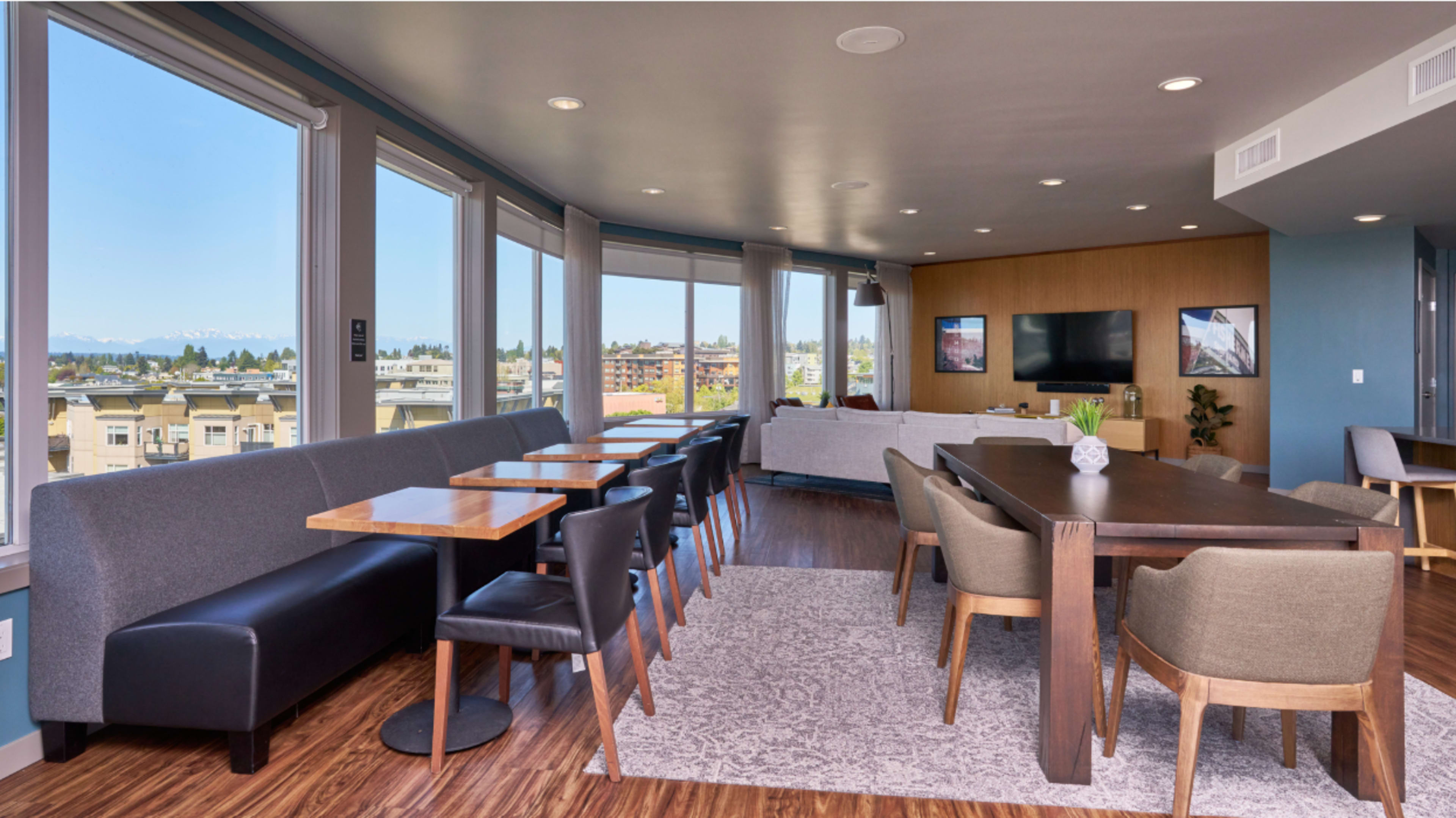 The image shows a modern living space with large windows, featuring a dining area with multiple tables and chairs, a sofa arrangement, and a television against a wall.