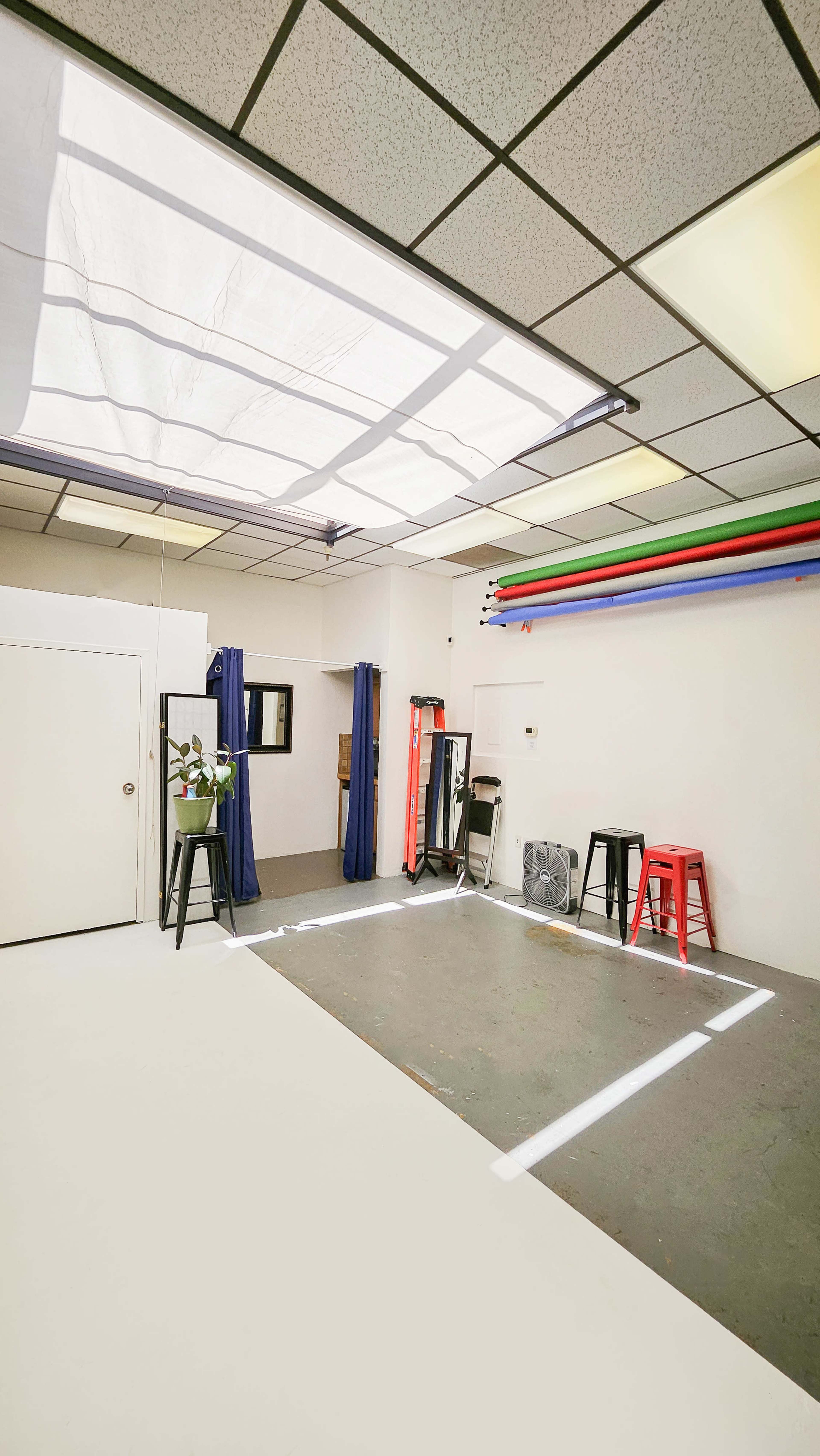 A spacious room with a white floor, a skylight, and colorful strips along the ceiling, featuring red and black stools, a fan, and curtains along the walls.