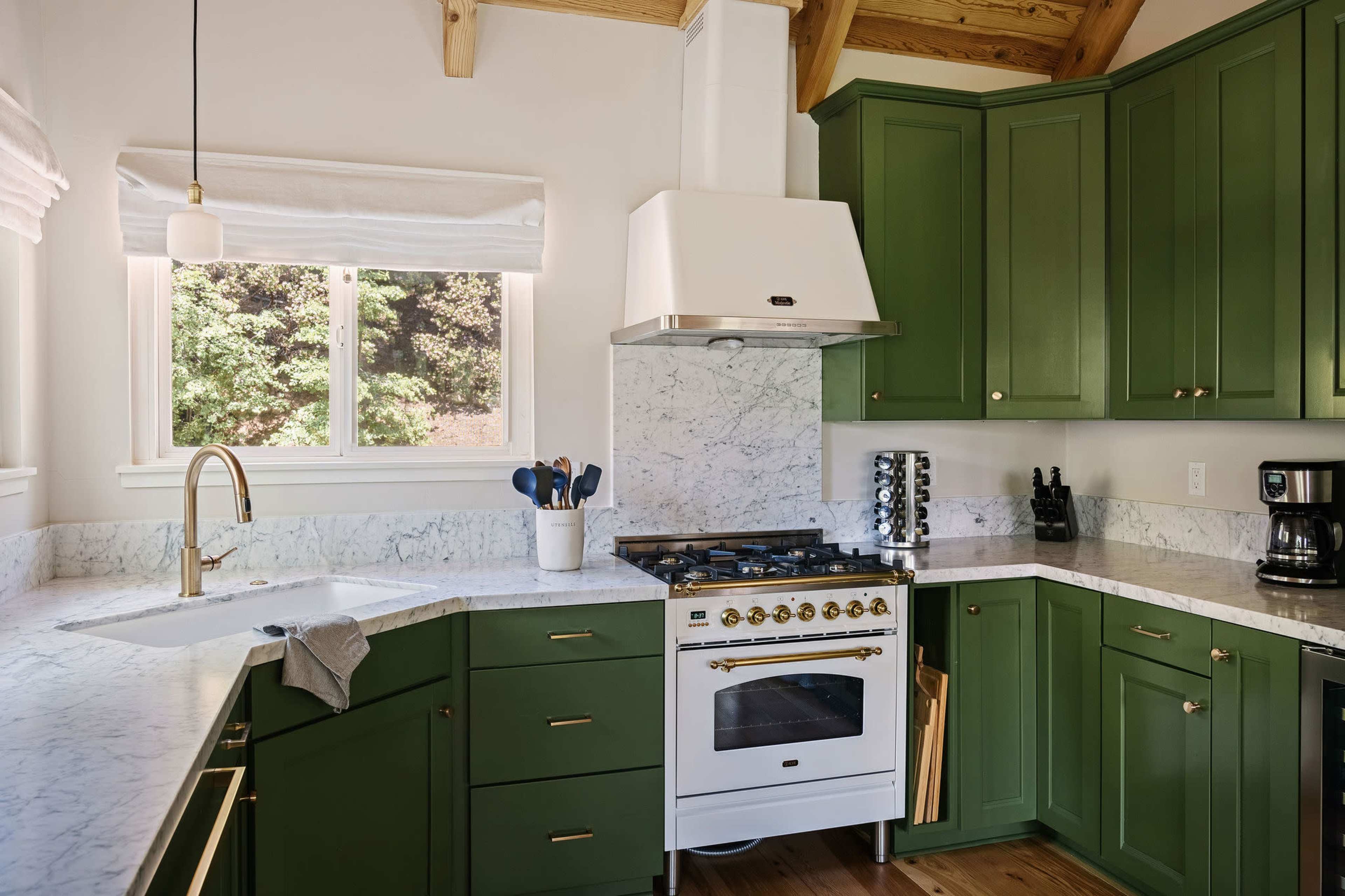 A modern kitchen with green cabinetry, a marble countertop, and a gas stove under a hood, illuminated by natural light from a window.