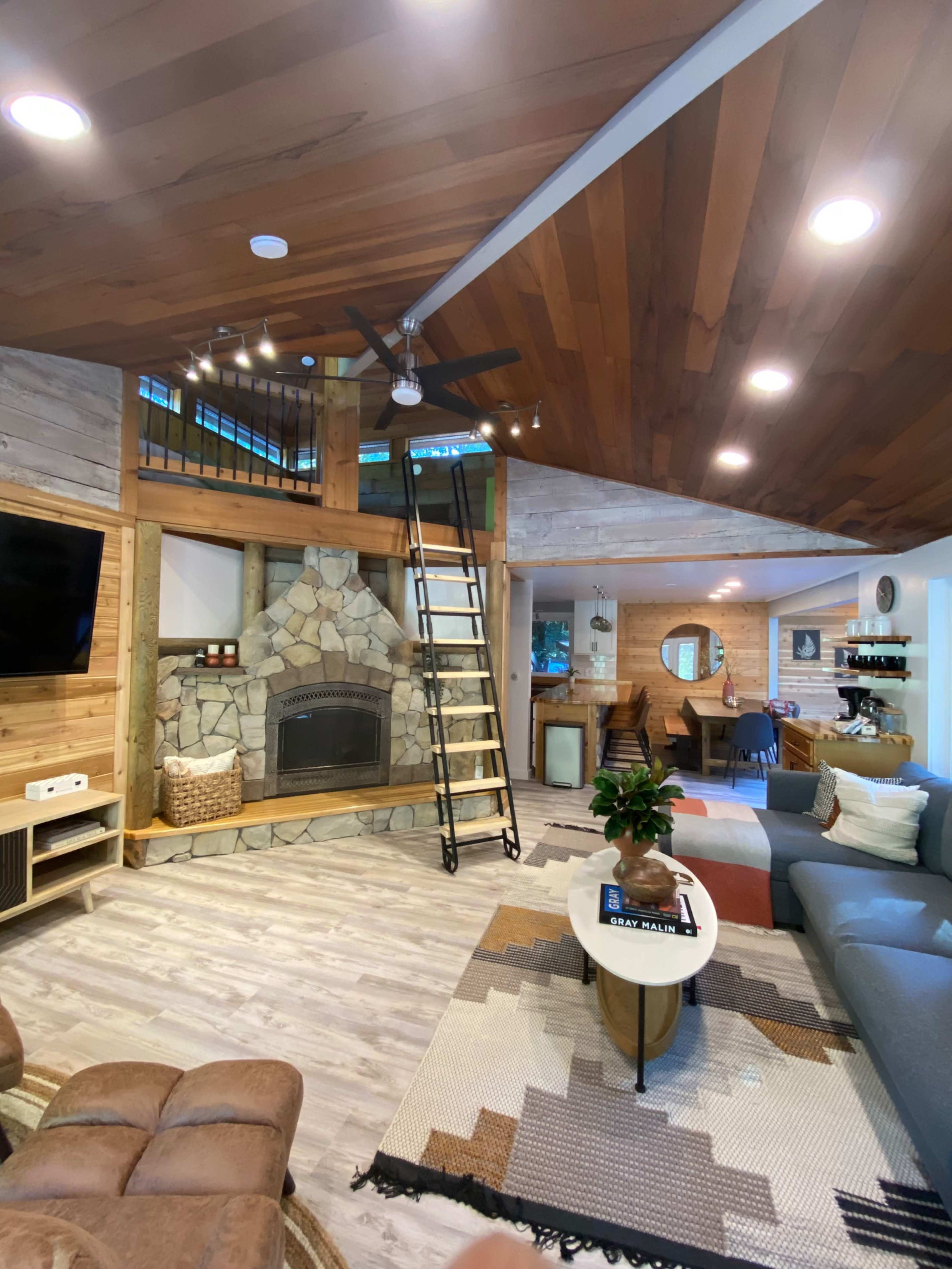 A living room features a stone fireplace, a wooden ceiling, a ladder leading to a loft, and modern furnishings.