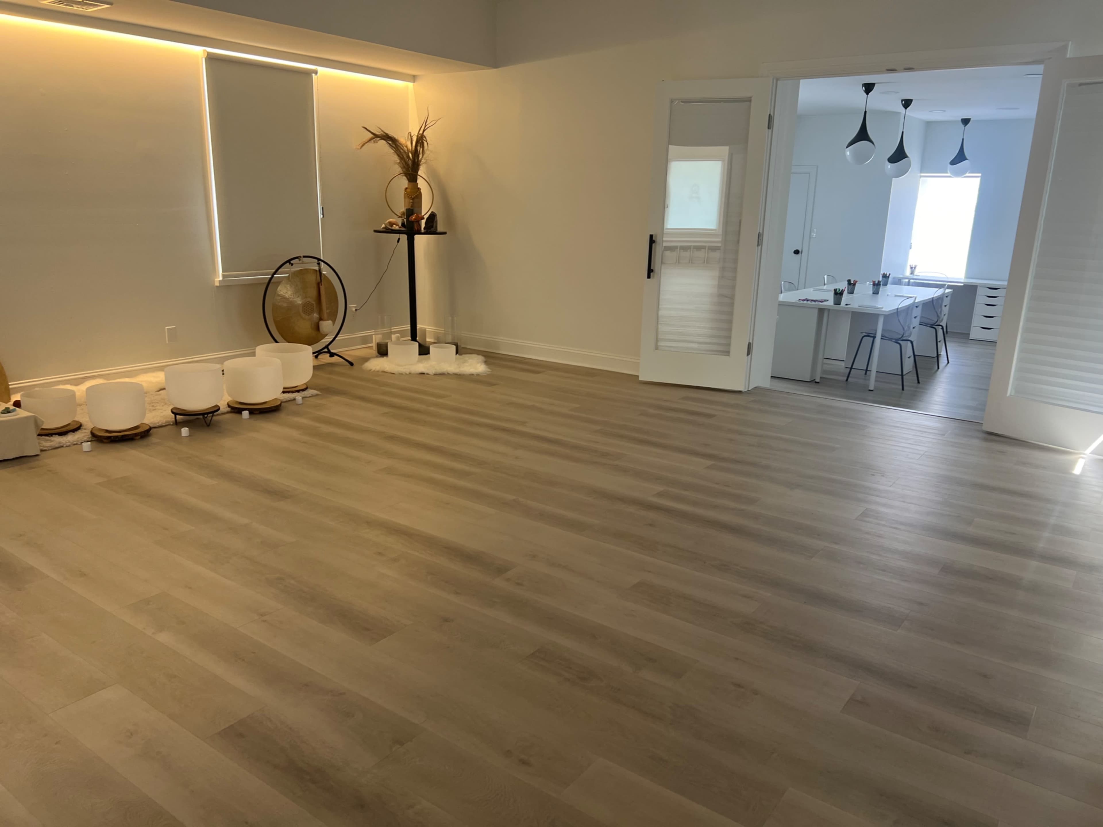 A spacious, empty room with light wooden floors, a simple table set for dining in the background, and decor items on a stand to the left.